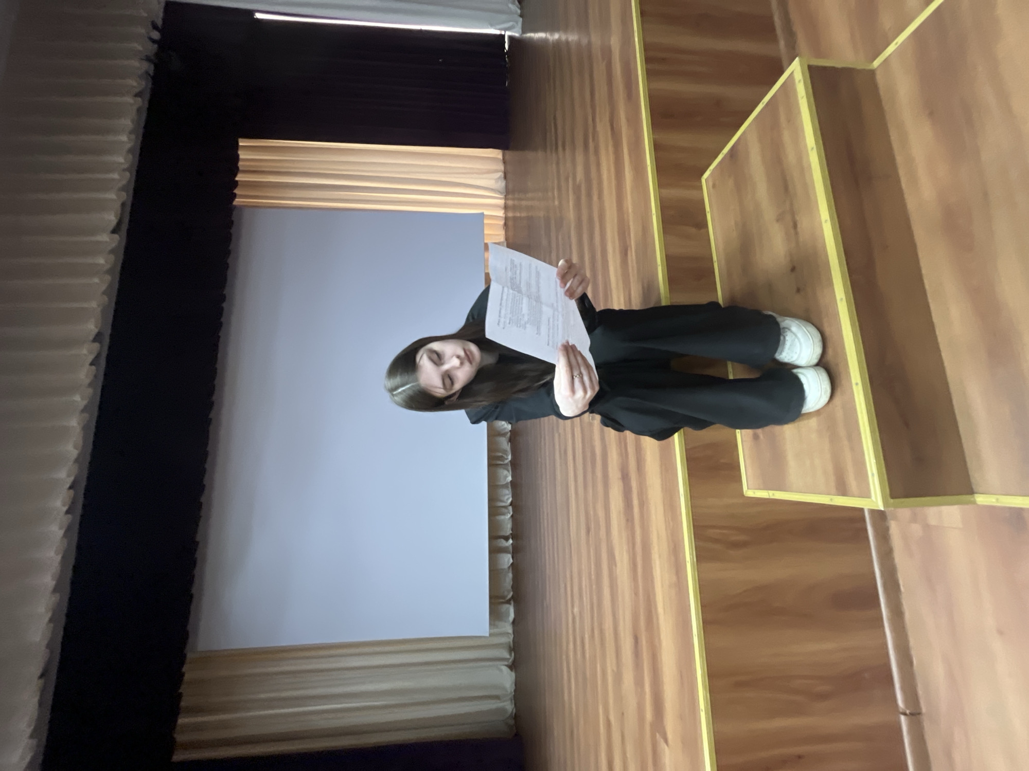 A young woman is standing on a wooden stage. She is holding a piece of paper. The stage has a backdrop of a white screen with gold curtains, and a black curtain on the left. The location is Saint Petersburg, Russia, and the woman is likely at a theatre or auditorium for a performance or event.