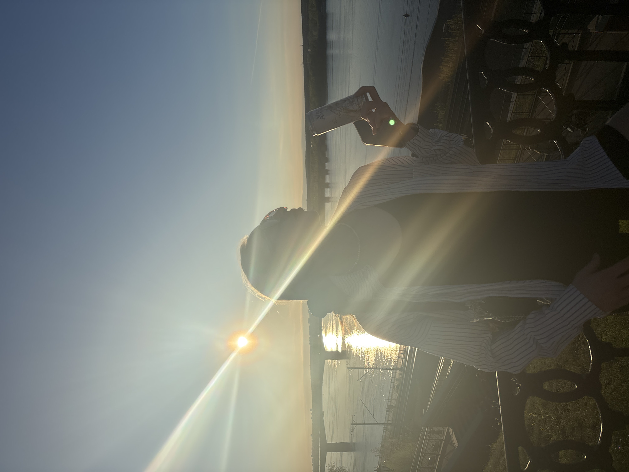 A person sits on a bench in Perm, Russia. The person is holding a can, potentially a beverage. The sun is bright, creating glare in the image. The Kama River is visible in the background. Railway lines are also present near the river.