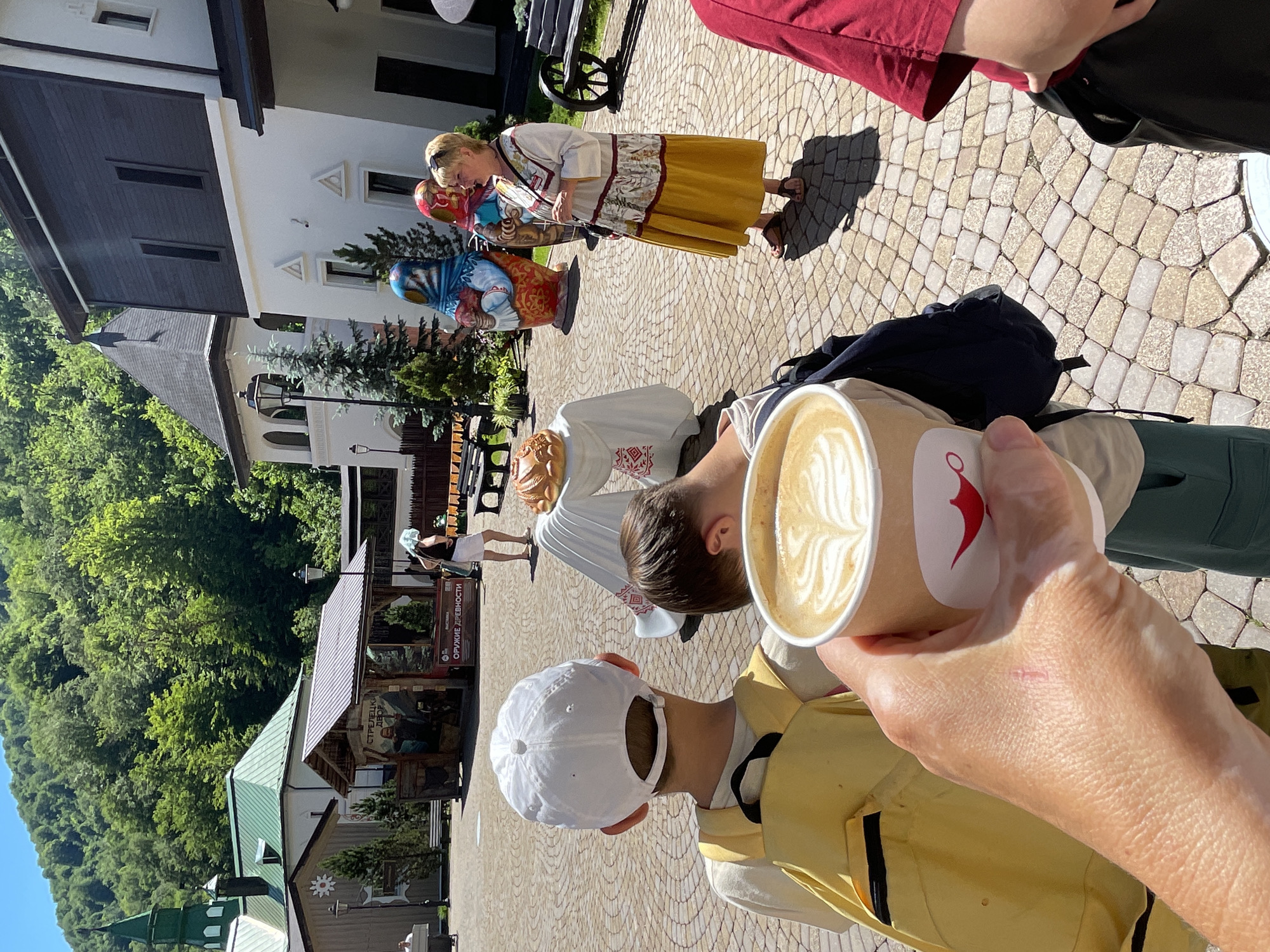 In the city of Estosadok, Russia, a paved courtyard is depicted on a sunny day. In the foreground, a person's hand holds a coffee cup with latte art, obscuring the view of two young boys in the middle ground. One boy, facing away, wears a white cap and a yellow backpack. The other boy, also facing away, has dark hair and is wearing a backpack. In the background, a woman in traditional Russian attire stands near large Matryoshka dolls and stalls. Buildings with pitched roofs and greenery are visible behind the stalls and dolls, and a mountain covered in trees looms in the distance.
