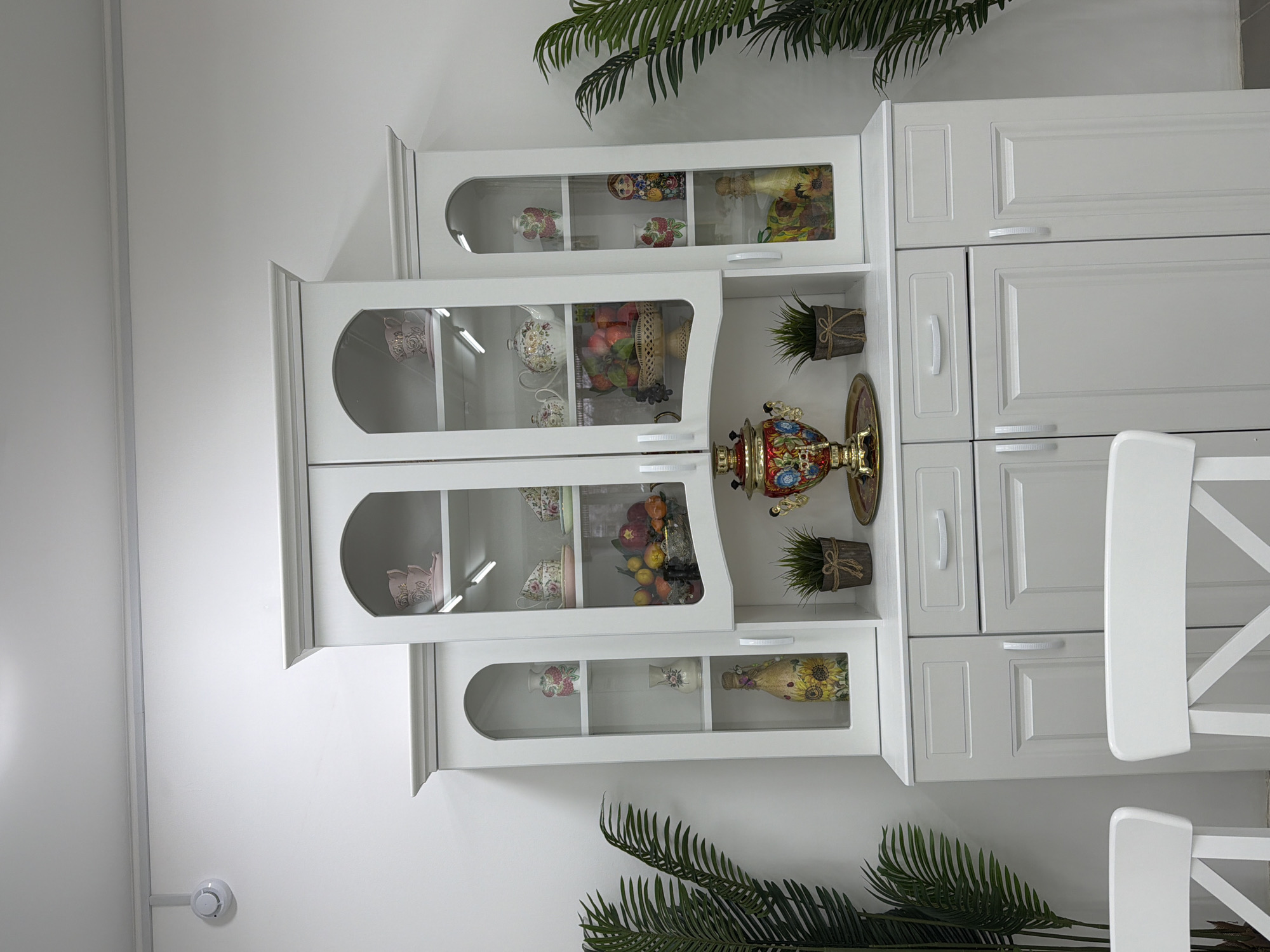 The image captures a clean, well-lit interior scene, likely a kitchen or dining area in Votkinsk, Russia. The dominant features are white cabinetry and walls, creating a bright and modern aesthetic.
On the left side, a series of interconnected white display cabinets with glass doors showcase various decorative items. The uppermost cabinet features colorful matryoshka dolls and a patterned vase. Below this, another glass cabinet displays white teacups with floral motifs and a basket filled with artificial fruits. The next visible display section holds more ornamental teacups and vases.
To the right of these display cabinets, a section of white kitchen cabinets with solid, paneled doors and subtle silver handles is arranged vertically. Between the glass display units and the solid cabinets, an open shelf prominently displays a traditional, ornate samovar in red, gold, and blue, alongside a small potted artificial plant. Another small potted plant is situated on a lower shelf.
Partially visible on the right side of the frame are two white chairs with slatted backs, suggesting the presence of a dining table out of view. Large green artificial leafy plants are strategically placed, one atop the cabinet system and another near the bottom right, adding natural accents. A white smoke detector is mounted on the wall in the lower left corner of the image. The overall impression is one of neatness and decorative elegance, incorporating elements of traditional Russian culture. No visible text is present in the image.