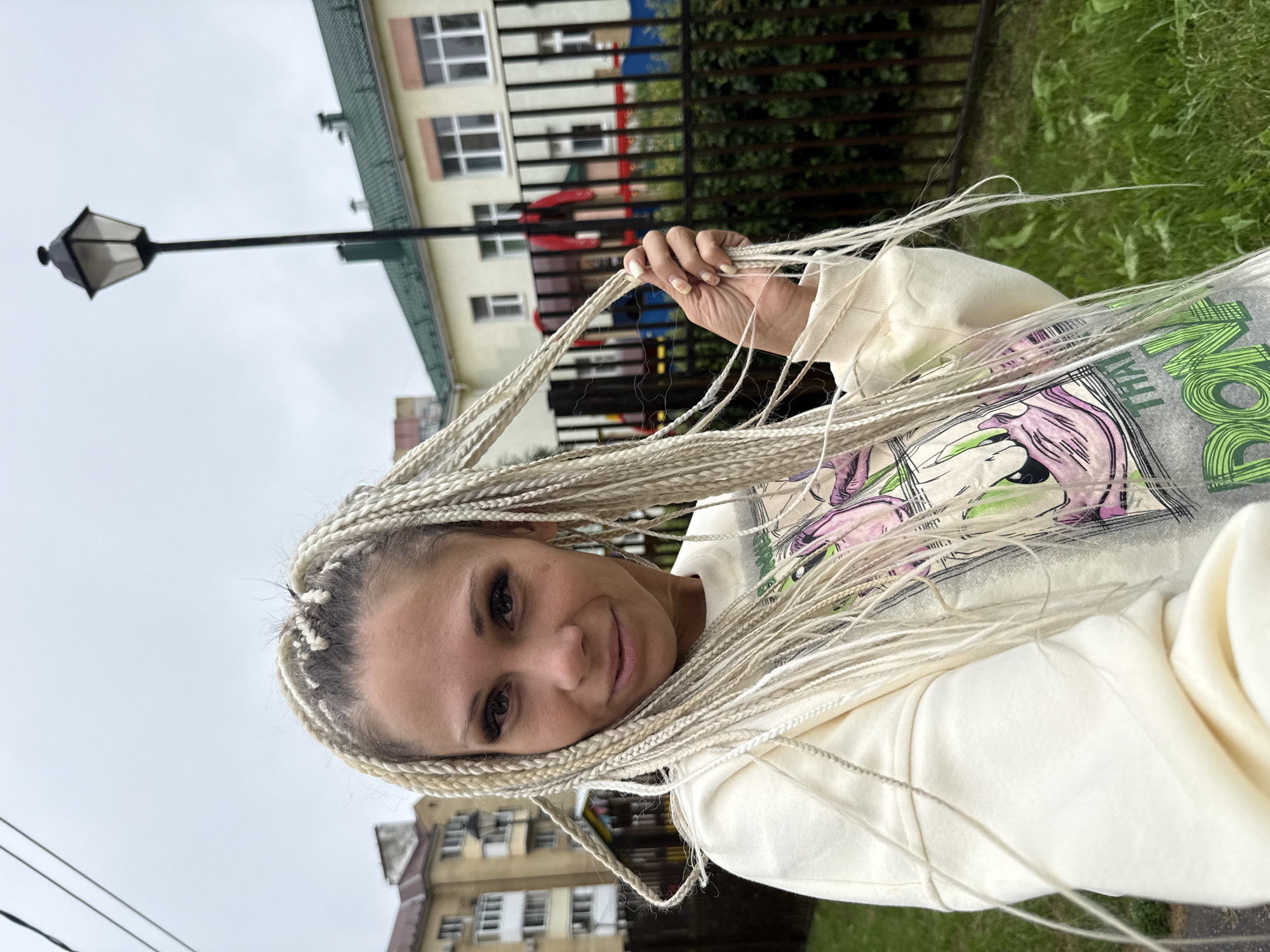 A medium shot shows a white woman with platinum blonde braided hair in an outdoor setting. She is looking at the camera with a slight smile. She is wearing a cream-colored sweatshirt with a graphic print on the front. Her right hand is raised, holding a section of her braids. The background includes a black metal fence, green grass, a building with a green roof, and a lamppost. The sky is overcast and gray. The location is identified as Rusanovo, Russia.