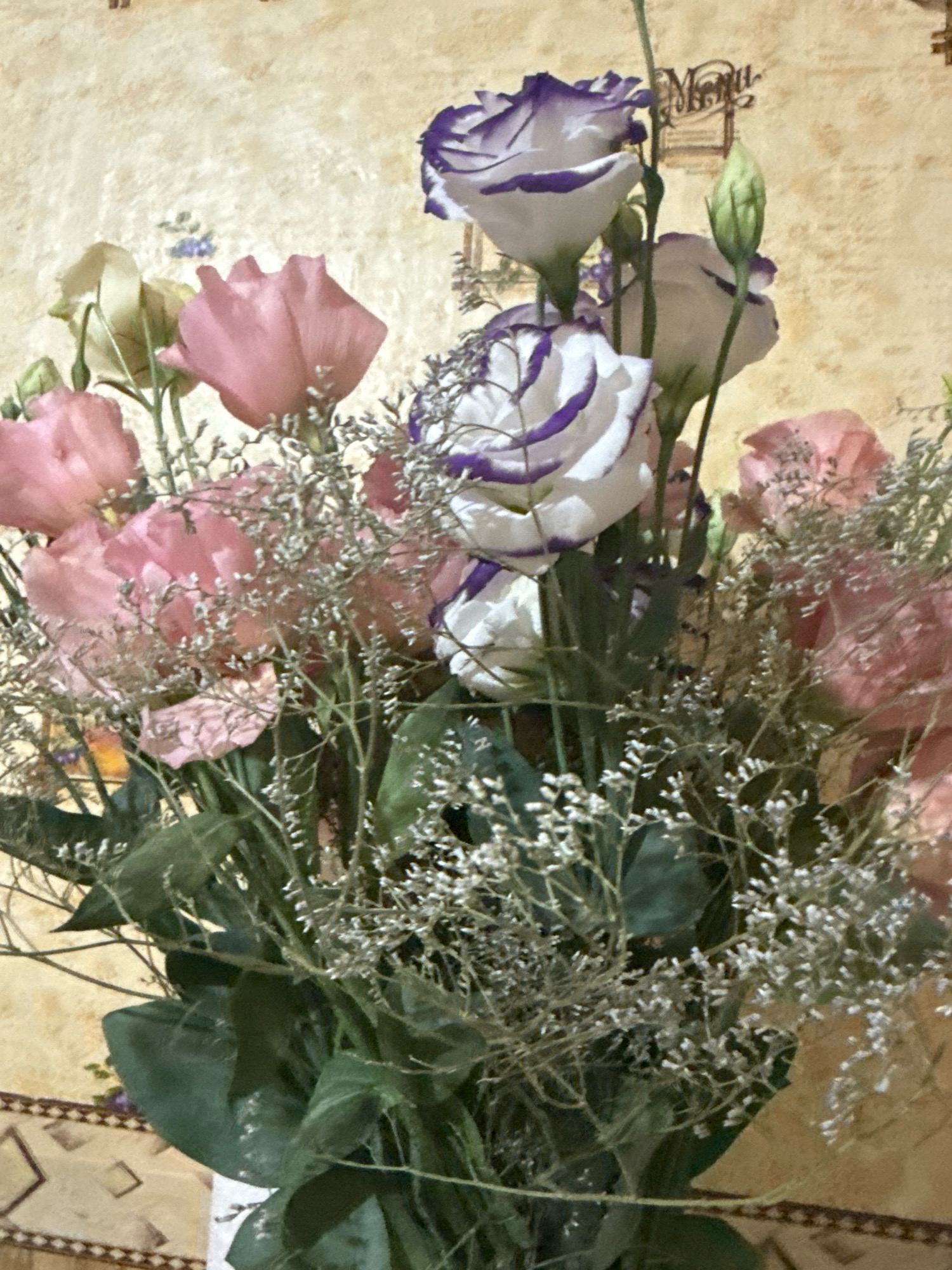 A close-up shot captures a bouquet of flowers, featuring a mix of pink lisianthus and white lisianthus with purple edges. The bouquet is filled with delicate sprigs of baby's breath. The flowers are arranged in a vase, the bottom of which is partially visible. The background is a textured, light-colored wall with what appears to be a menu or signage visible in the upper right quadrant. The lower part of the image shows a patterned surface, likely a table or floor, with diamond shapes and a decorative border. The lighting suggests an indoor setting. The city of Basarabeasca, Moldova, is not depicted in this image.