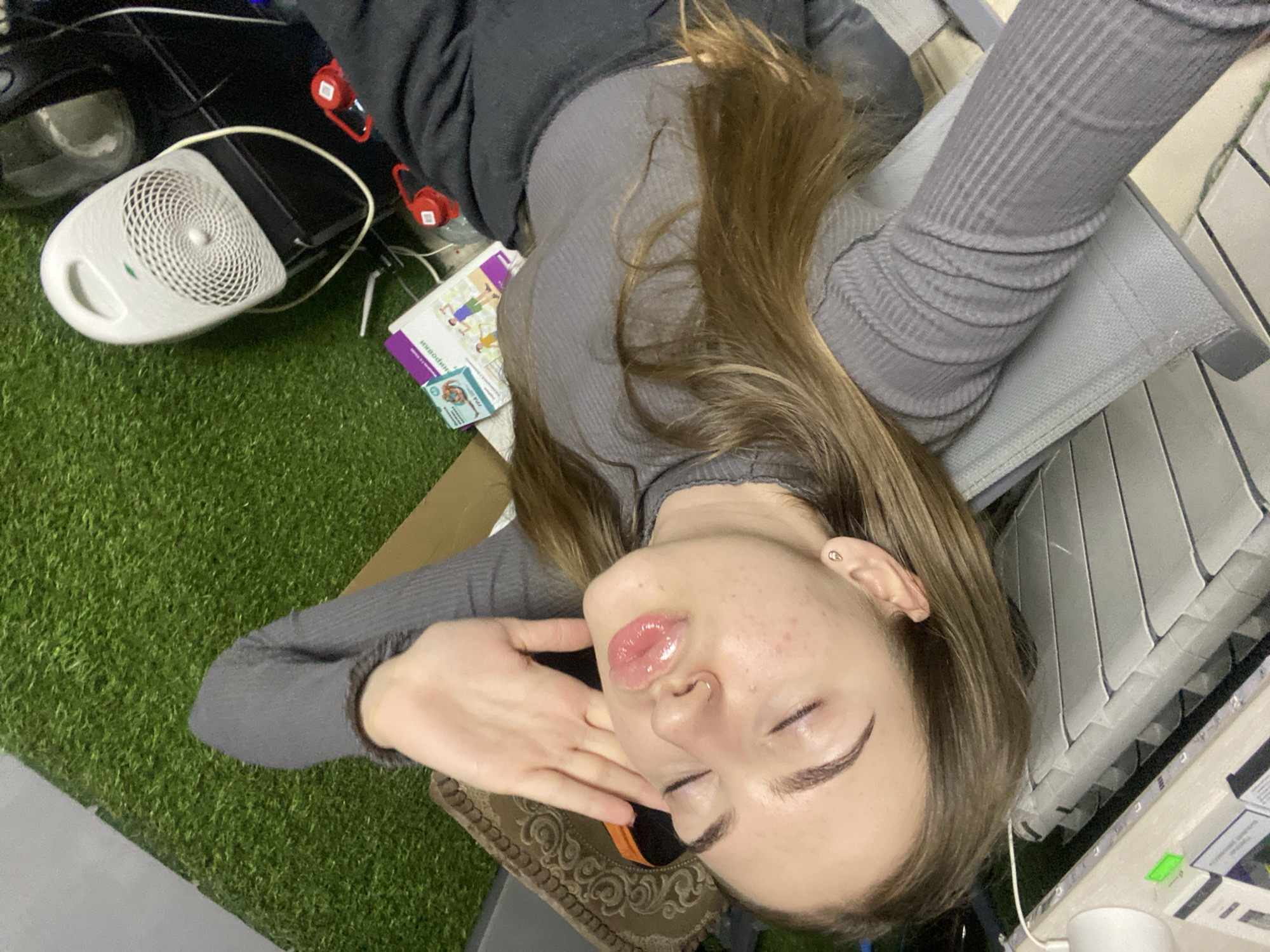A young woman with brown hair and fair skin is lying down and taking a selfie. She is wearing a grey long-sleeved shirt and black pants. Her eyes are closed, and her lips are pursed. Her right hand is raised to her cheek. The background is cluttered with various objects, including a white fan, a book, and a dark chair. The floor is covered with artificial grass. The scene appears to be indoors, in an urban setting in Dyakovo, Russia.