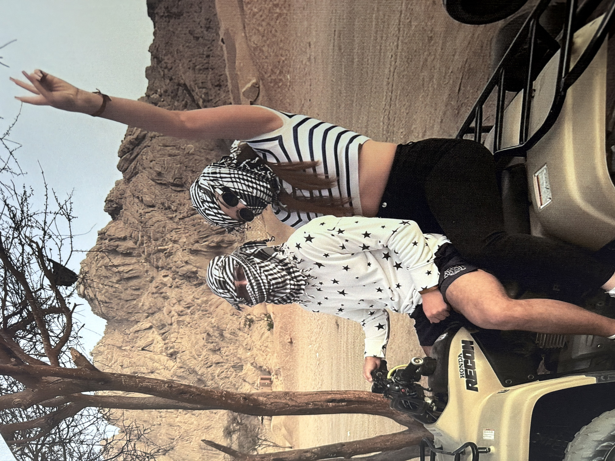 An image shows two individuals on an ATV in a desert environment. The person on the left is wearing a white long-sleeved shirt with a black star pattern and black shorts, with a black and white keffiyeh covering their head and face. They are seated on the ATV, holding the handlebars. The person on the right is wearing a black and white striped crop top and black pants, also with a black and white keffiyeh and sunglasses. This individual is seated behind the first person and has their right arm raised in a peace sign gesture. The ATV is a tan Honda Recon. The background consists of rocky, mountainous terrain and a sparse, leafless tree. The lighting suggests it is daytime. There is no visible connection to Strășeni, Moldova.