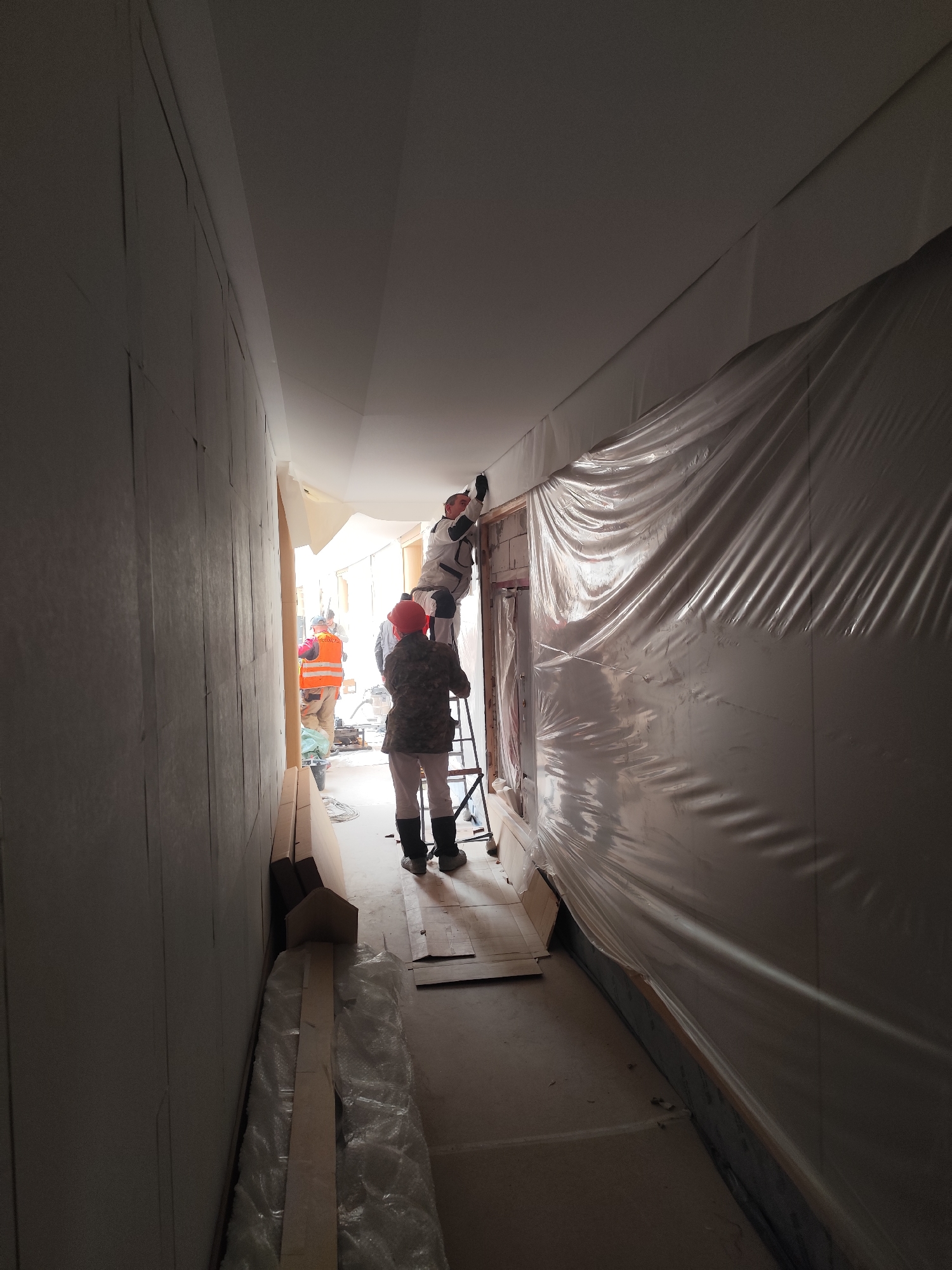 This image captures an indoor scene within a narrow hallway or corridor under renovation or construction. Three workers are present, engaged in interior finishing work.
In the mid-ground, a worker dressed in white overalls stands on a step ladder, actively working on the junction where the ceiling meets the right wall, possibly plastering or painting. Below, another worker wearing a red hard hat, a camouflage-patterned jacket, light pants, and dark boots stands near the base of the ladder, seemingly assisting or supervising. Further down the corridor, in the background, a third worker is visible wearing an orange high-visibility vest.
The left wall of the hallway is covered with panels, possibly plasterboard, showing visible taped seams. The right wall is largely draped with clear plastic sheeting, extending upwards to cover parts of the ceiling, indicating protection against dust or paint during ongoing work. The ceiling itself appears light-colored and newly prepared. The flooring is an unfinished concrete or subfloor. Along the base of the left wall, wooden planks or beams wrapped in plastic and cardboard pieces are stacked.
The lighting in the corridor appears to originate from the far end, illuminating the background and the work area, creating a sense of depth. The atmosphere suggests focused activity within an active construction or renovation site. No discernible text is visible on any objects or clothing.