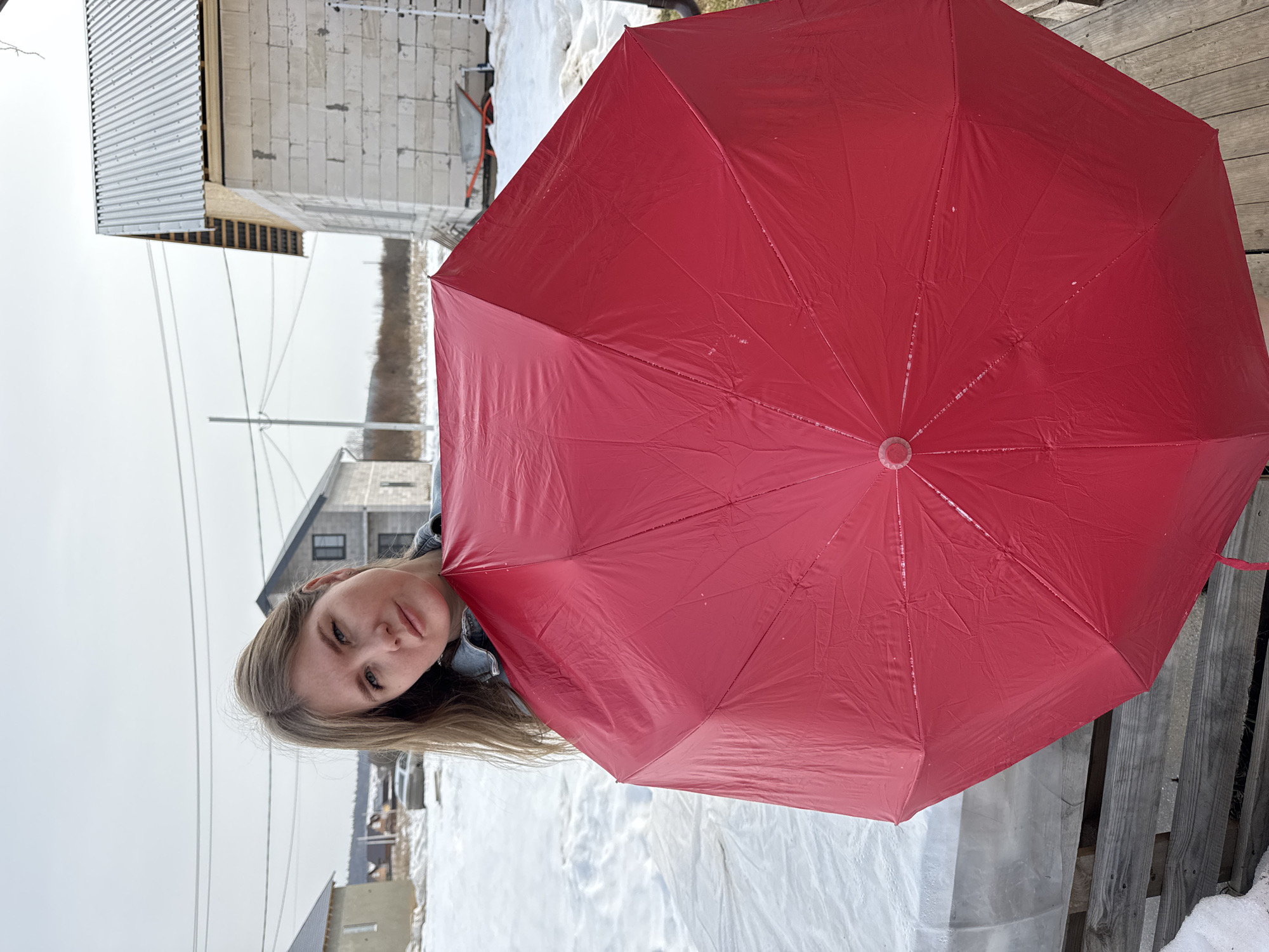 The image depicts a woman partially visible on the left side of the frame, peeking out from behind a large, bright red umbrella that dominates the right and central portions of the scene. The image is taken outdoors on a cloudy, overcast day, with snow covering the ground.
The woman has long, straight, light brown or blonde hair that falls over her shoulders. She is looking directly at the camera with a neutral expression. She appears to be wearing a dark-colored top, possibly a denim jacket, under the umbrella.
The red umbrella is open and held up, facing towards the viewer. Its surface appears slightly wrinkled and has several small white specks or water droplets on it, suggesting it might be damp or have recently been used in precipitation. The center cap of the umbrella is visible, showing its radial structure.
The background consists of a snowy landscape and several buildings. On the far left, a structure with a corrugated metal roof and unfinished light-colored block walls is visible, possibly a building under construction. Further back, toward the center-left, another building, possibly a house, with dark windows and a dark roof can be seen. Utility lines or power lines stretch horizontally across the middle distance of the left side of the frame, silhouetted against the bright, overcast sky. The ground is covered in a layer of snow, with some areas featuring white plastic sheeting or tarps covering the snow. On the far right, behind the umbrella, a wooden fence or structure with horizontal planks is partially visible.
The overall scene suggests a cold weather setting, likely winter or early spring. The sky is bright but uniformly gray, indicating a lack of direct sunlight. No discernible text is visible in the image.