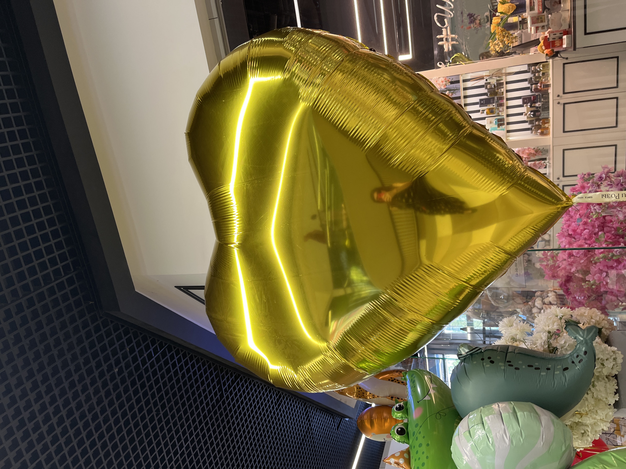 A large, reflective, gold, heart-shaped balloon dominates the foreground of the image. The balloon is a solid, metallic gold color, with visible creases and lines from its inflated state. Its surface reflects the surrounding environment, including a distorted image of a person holding a phone, presumably taking the photo, wearing sunglasses and a dark top. Behind the balloon, to the left, a cluster of other balloons is partially visible. These include a green alligator-shaped balloon with large eyes, a round orange balloon, and a white and green striped balloon. Further in the background, there is a display of shelves stocked with various bottles, likely perfumes or other small retail items, against a black and white striped wall. Pink and white floral arrangements are also visible in the mid-ground and background. The ceiling above is a dark, grid-like structure. The overall setting appears to be an indoor retail space, possibly a party supply store or a gift shop. The lighting suggests an interior setting with artificial lights contributing to the reflections on the balloon. The location is unknown, as is the city.