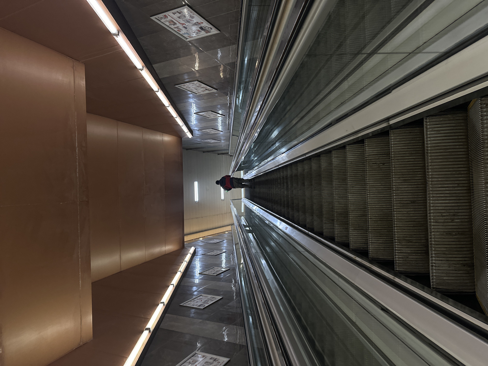 An indoor, vertically oriented long-exposure image depicts a subterranean passage, likely a metro station in Sofia, Bulgaria. The composition is dominated by a long escalator on the right side, which appears to be descending. The escalator features metallic steps and a transparent or reflective balustrade, with the continuous handrail visible along its length.
A lone individual, dressed in a dark coat with a red backpack or detailing, is positioned on the escalator about halfway down, facing away from the viewer.
To the left of the escalator, a polished, dark-tiled floor reflects the bright linear fluorescent lights that run along the base of the adjacent wall and across the ceiling. Scattered along the floor and mounted on the ceiling are several rectangular framed displays, possibly posters or information panels, though their specific content is unreadable due to distance and resolution.
The left side of the passage is lined with smooth, matte brown or reddish-brown wall panels, illuminated by the continuous strip lighting. The overall aesthetic is clean and modern, characteristic of public transportation infrastructure, with strong lines and artificial lighting creating a sense of depth and movement.