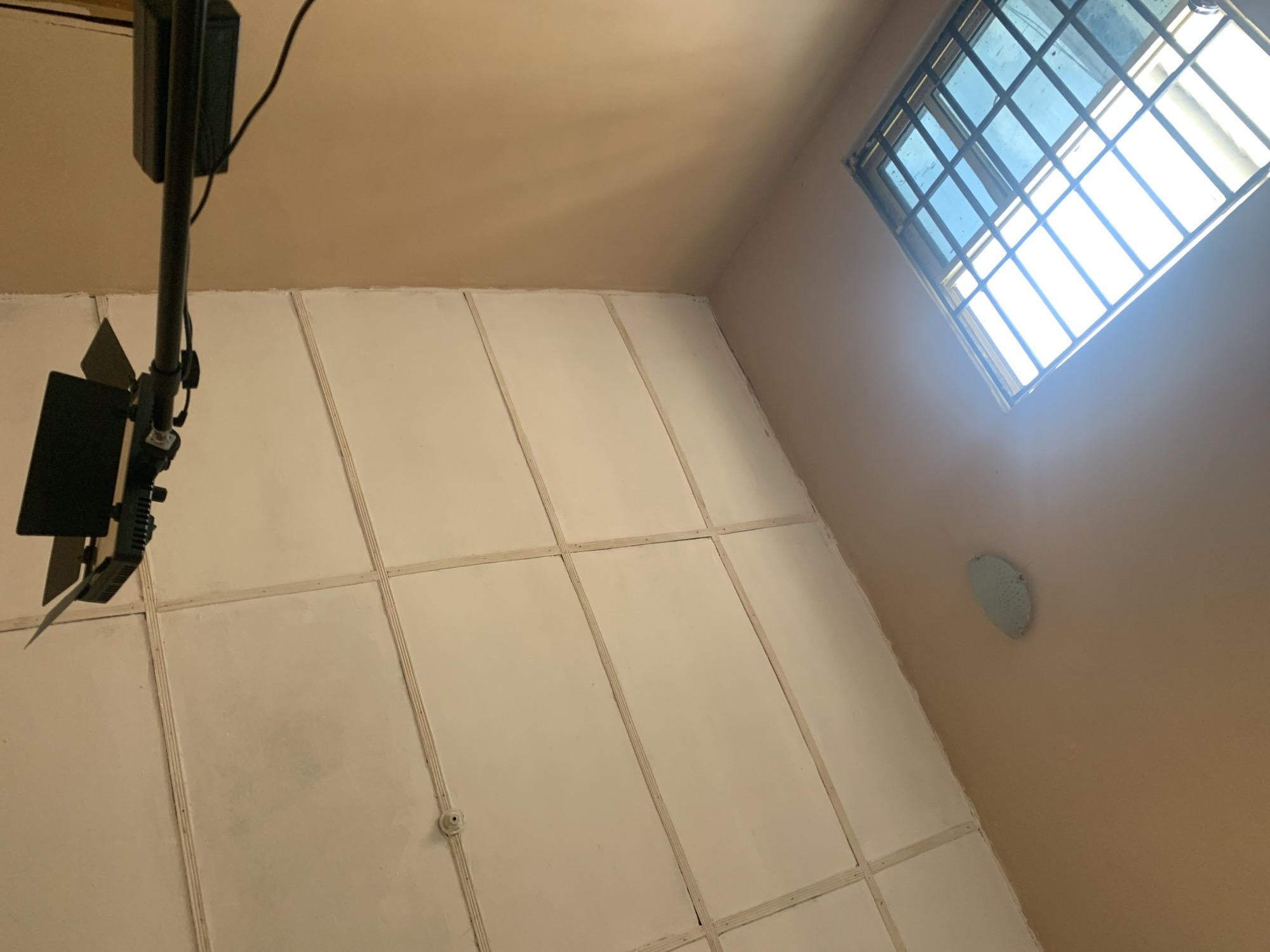 A ceiling light fixture is mounted to a black pole, extending horizontally from the right side of the frame. The fixture has black barn doors. Below the fixture, a section of the white, paneled ceiling is visible. To the left of the ceiling, a small, circular, white device is mounted. Further down on the tan wall, a blue-tinted window with a black metal grate is visible, with bright light shining through it.
