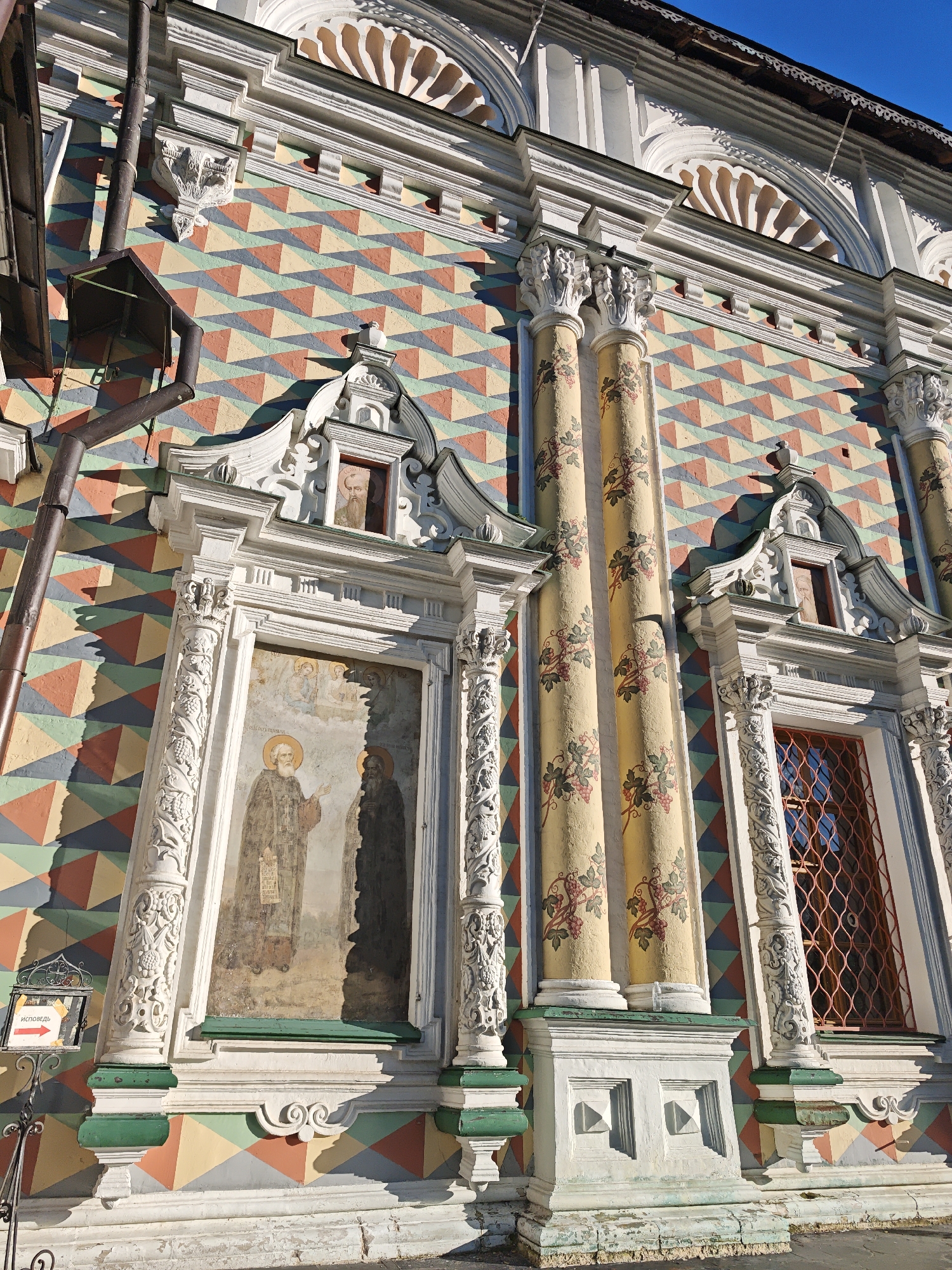 The image shows a detailed view of the exterior of a building with a highly decorative facade. The wall is adorned with a geometric pattern of colorful triangles in shades of orange, yellow, green, and grey. Two fluted columns, painted yellow and decorated with painted vines and clusters of grapes, stand prominently in the foreground. These columns support ornate white capitals. Between the columns are two recessed niches containing religious icons. The left niche has a larger icon depicting two figures, one with a halo. The right niche contains a smaller icon of a bearded man with a halo. Above these niches are architectural elements featuring white stucco work and decorative arches. A brown downspout runs vertically along the left side of the building. A metal sign with Cyrillic text and a red arrow pointing right is attached to a metal stand to the left of the left icon. The building appears to be made of stone and is bathed in bright sunlight, casting shadows on the patterned wall. The overall impression is one of ornate and historical religious architecture.