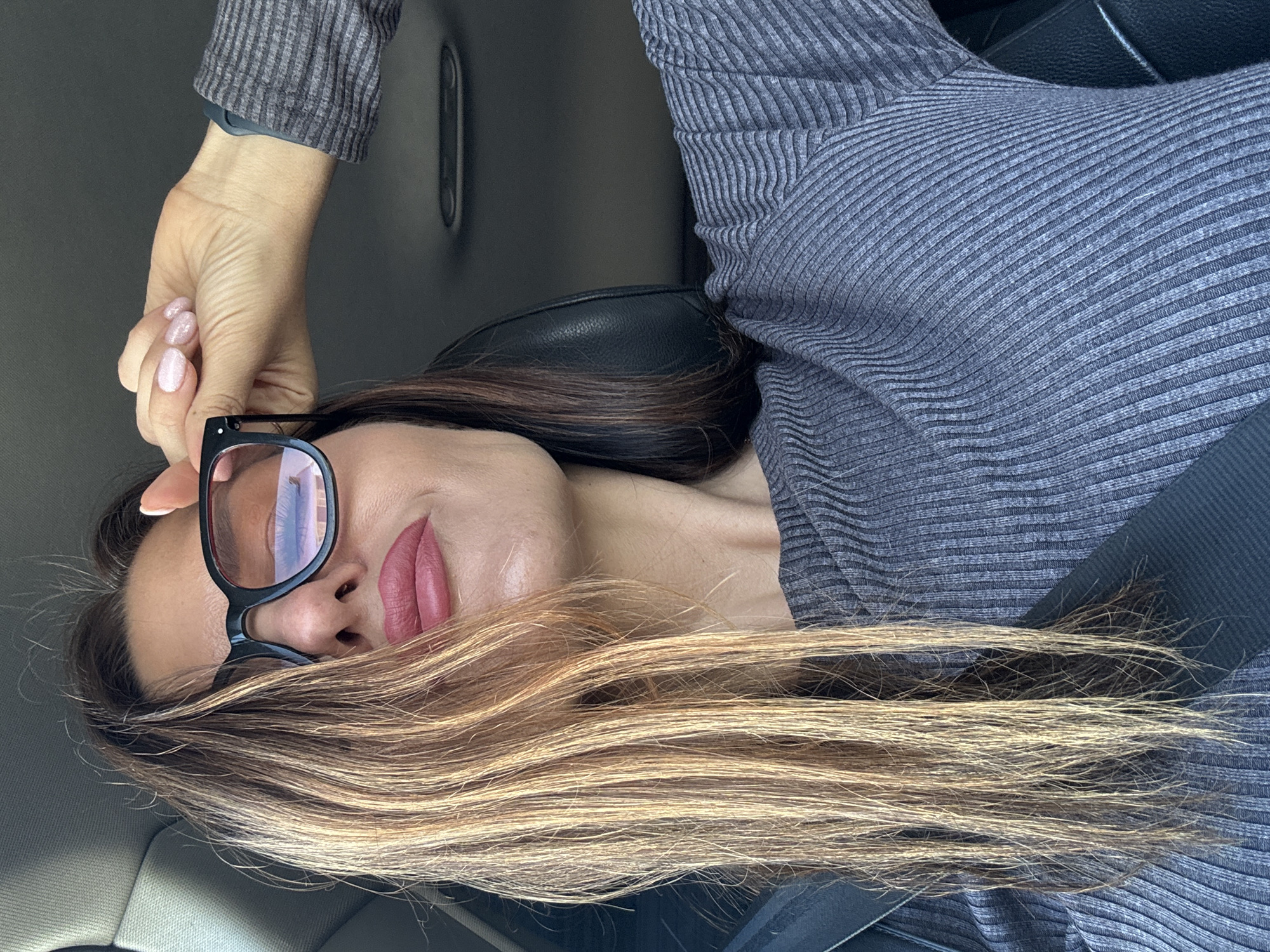 A woman wearing a gray ribbed top and black-framed glasses is shown inside a vehicle. She is wearing a seatbelt. Her hair is partially obscuring her face. The location is unknown and is not discernible from the image.