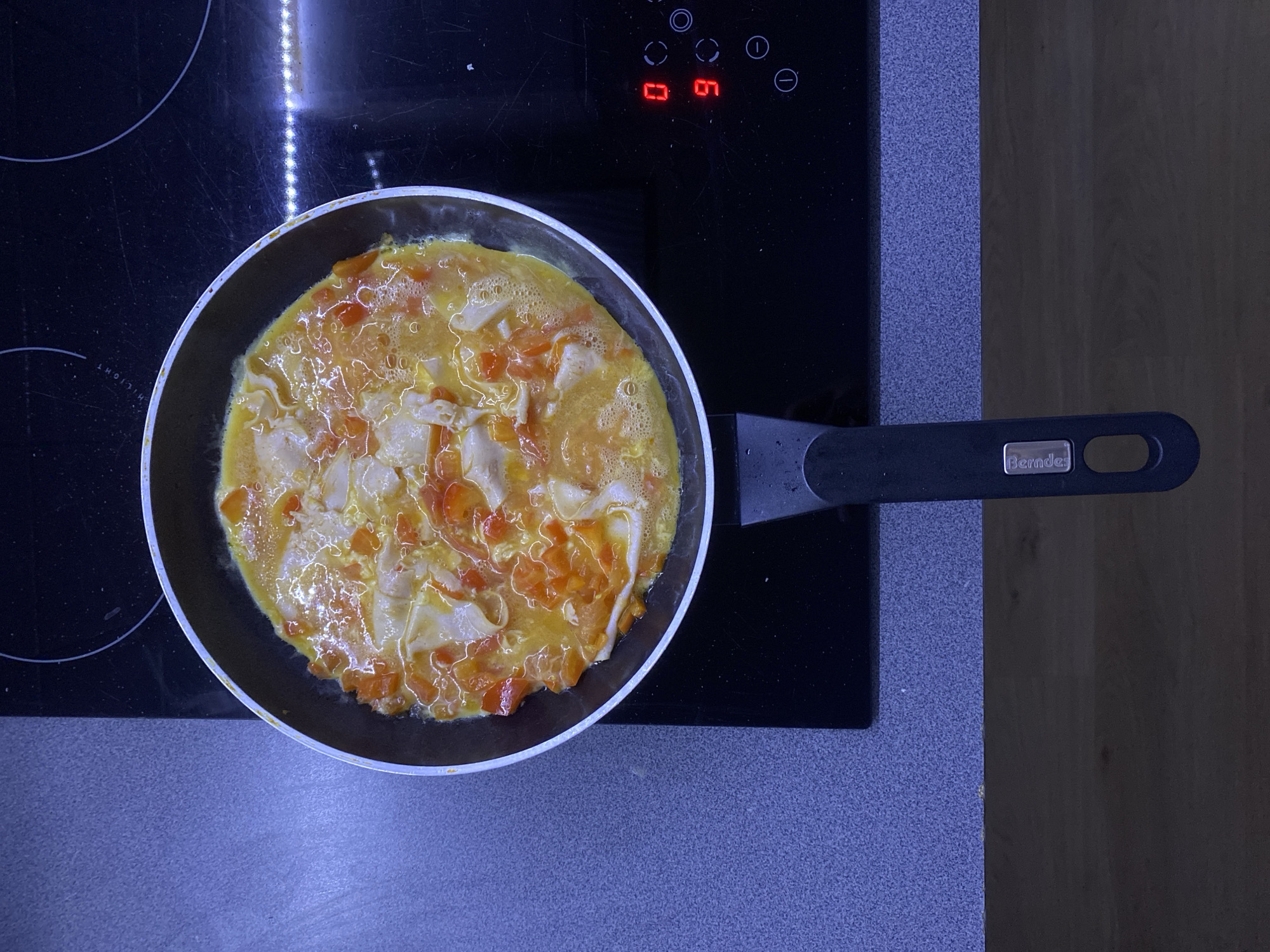 A high-angle shot shows an omelet cooking in a black frying pan on a black induction stovetop. The omelet is a mix of scrambled eggs and chopped tomatoes, with some lighter-colored pieces that could be cheese or chicken. The frying pan has a black handle with a silver nameplate that reads "Berndes." The stovetop has touch controls, with a red digital display showing "06." The scene is dimly lit, suggesting it may be nighttime or indoors. The background shows a textured surface that could be a countertop and a wooden surface, indicating a kitchen setting. The image appears to be taken in a home environment. No people are visible in the frame.