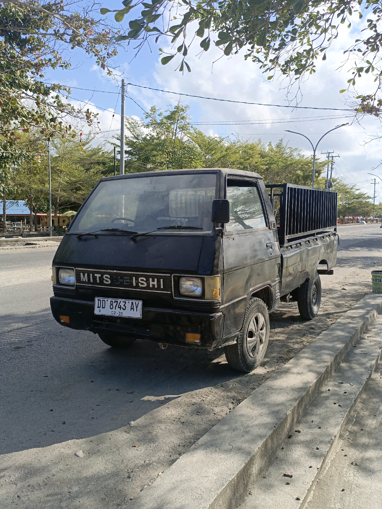 A black Mitsubishi pickup truck is parked on the side of a road in Bulukumba, Indonesia. The truck has a license plate that reads "DD 8743 AY". The truck is facing the viewer and is angled slightly to the left. The truck bed is empty and has a black metal cage on top. The truck appears to be old and somewhat worn.
The road is paved and there is a concrete curb on the right side of the image. There are trees and buildings in the background. The sky is blue with some clouds. The lighting suggests it is daytime. The environment appears to be a moderately developed area with some vegetation.