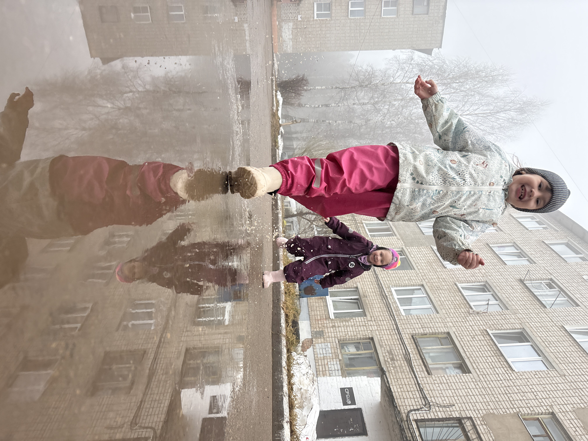 A photograph taken in what appears to be Chaikovsky, Russia, shows two young girls playing near a puddle that reflects the surrounding architecture and bare trees. The girl closest to the viewer wears a light-colored patterned jacket, magenta pants, and a striped beanie. She has her arms outstretched and appears to be smiling. The second girl, standing closer to the puddle, wears a purple snowsuit with a pink beanie, also with her arms out. The puddle surface is reflective, showing the girls and the nearby building, a large brick structure with rows of windows. The scene suggests a cold or overcast day, with a general gray hue and some visible fog.