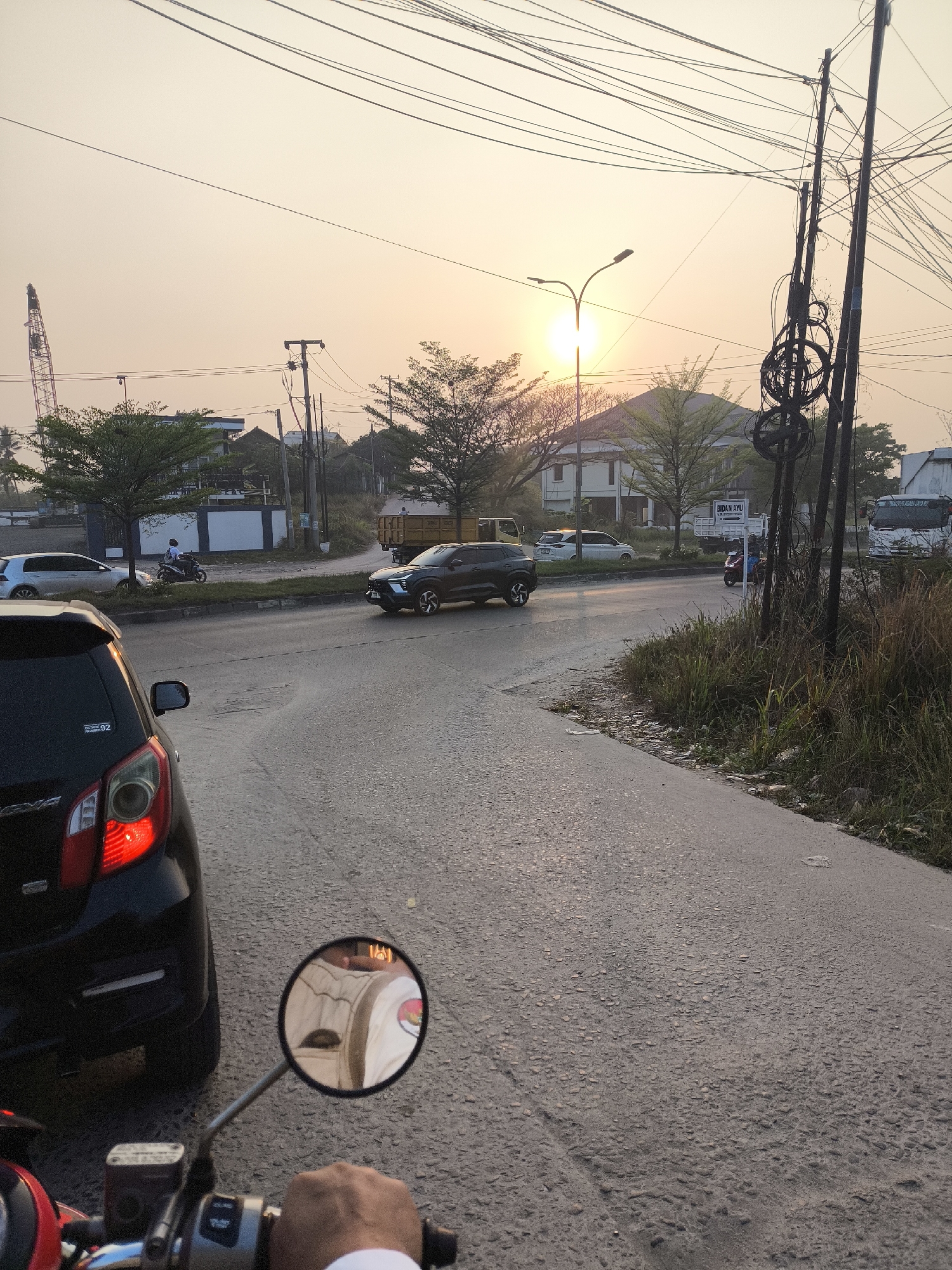 A personal perspective shot taken from a motorcycle shows a street scene during sunset in Cilegon, Indonesia. The road is paved with asphalt and shows signs of wear. In the foreground, the handlebars of a motorcycle are visible, along with a hand gripping them. A rearview mirror attached to the handlebar reflects the rider's white shirt and part of their face.
To the left of the road, a black car is parked, and further down the road, a silver car is driving. On the right side of the road, tall grass and weeds grow along the embankment. In the middle ground, several vehicles are on the road, including a dark SUV making a turn and a yellow truck. In the background, buildings, trees, and utility poles with a complex network of wires are visible. The sky is filled with the warm hues of a sunset, with the sun partially obscured by trees. A construction crane is visible in the far distance. The scene suggests a typical evening commute in an urban or suburban environment.