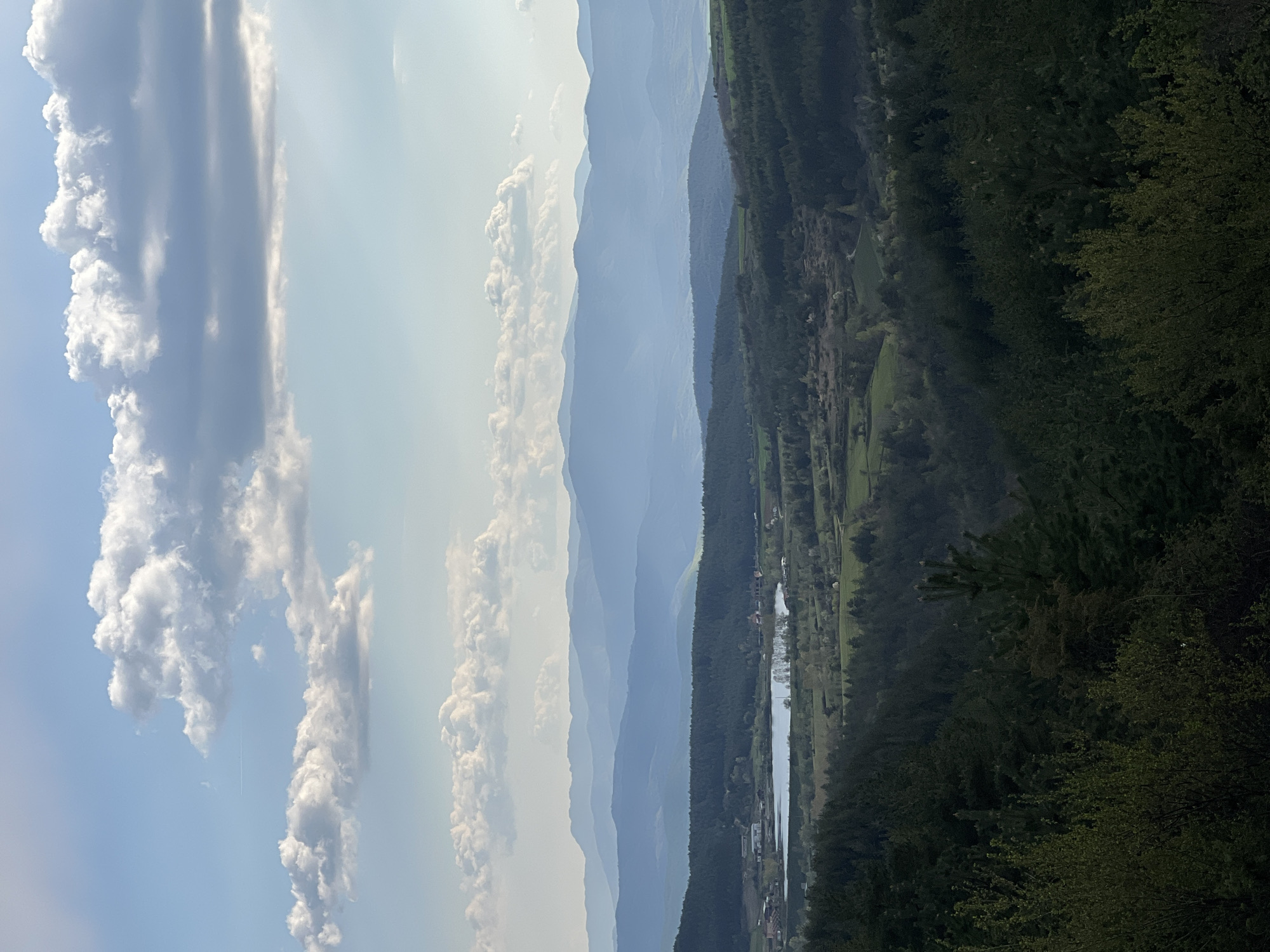 The image depicts a landscape with layered mountains fading into the horizon. Cumulus clouds are present in the sky. A body of water, likely a river or lake, winds through a valley surrounded by dense forest. There is evidence of human settlement or development along the water's edge. While the image generally aligns with typical Bulgarian mountain terrain, a precise determination of location within Kribul requires additional contextual information from someone on the ground or with local expertise. Without explicit confirmation, precise identification is impossible.