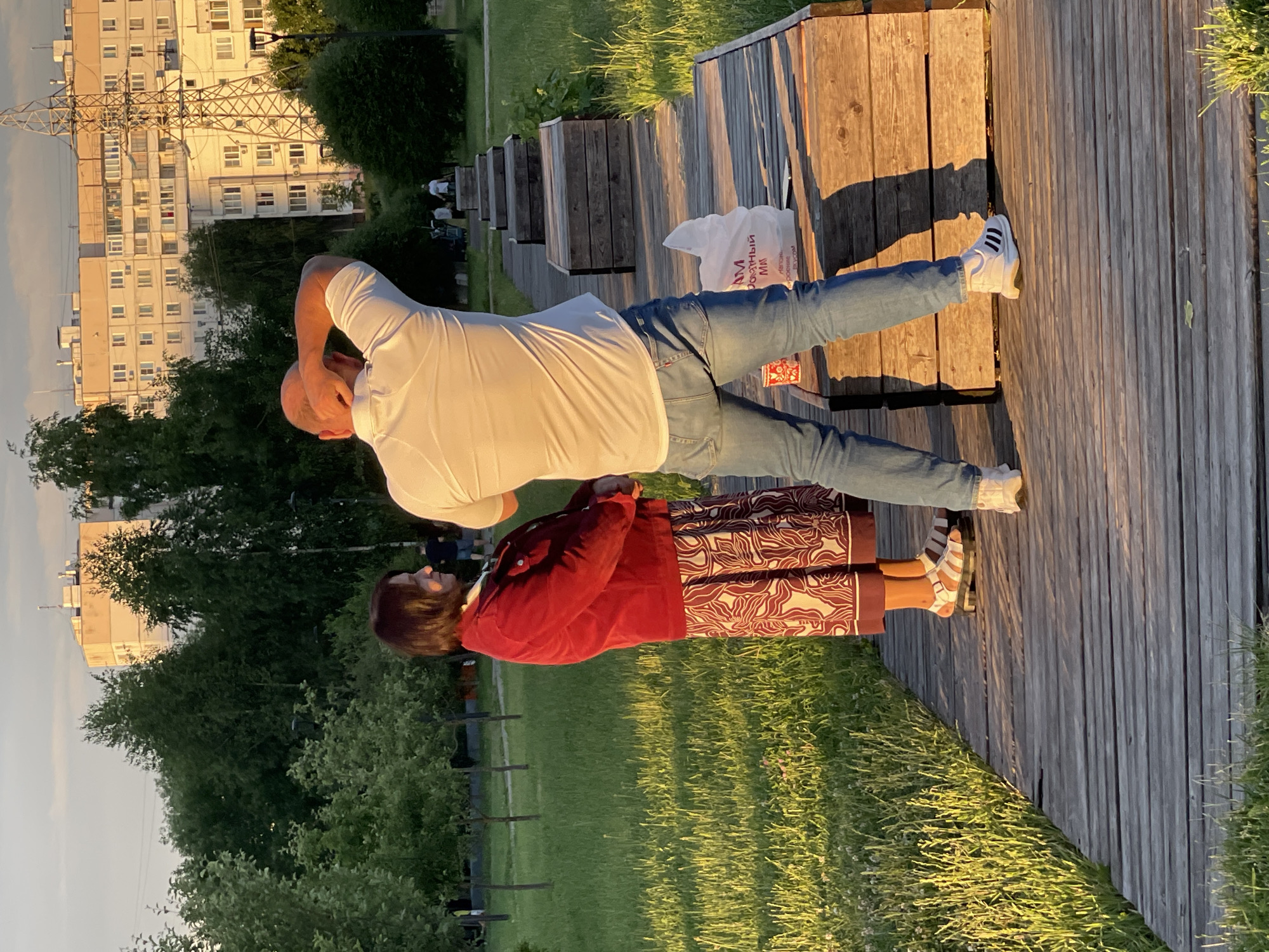 In Moscow, Russia, two individuals stand on a wooden boardwalk in a park setting. The woman, facing left, wears a red jacket and a patterned skirt. The man, with his back to the camera, is in a white polo shirt and blue jeans, holding his hand to the back of his head. A white plastic bag rests on the boardwalk near his feet. In the background, trees line the park, and a tall, light-colored residential building complex is visible along with a large metal pylon. The sun is low in the sky, casting long shadows and illuminating the grass with a golden light.