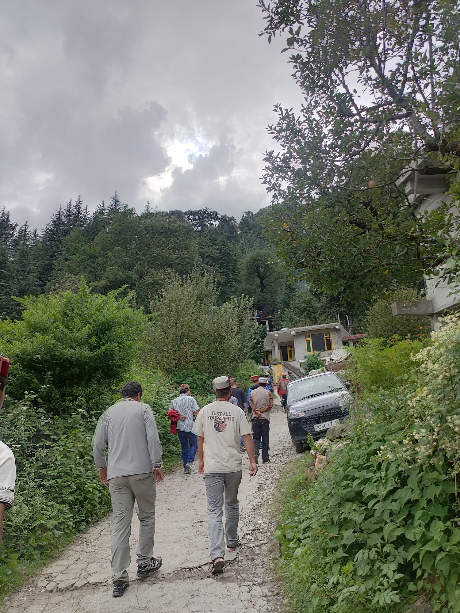 This is a photograph taken in Burwa, India, showing a group of people walking along a rough, unpaved road. The environment is rural and mountainous, with dense green vegetation and trees lining the road. The sky is overcast with thick clouds, suggesting a possibly rainy or humid day.
In the foreground, three men are walking away from the camera on the road. The man on the left wears a light grey long-sleeved shirt and grey trousers. The man in the middle wears a white t-shirt with text and graphics, grey trousers, and a cap. The man on the right wears a blue and white striped shirt, dark trousers, and a red jacket is slung over his shoulder.
Further up the road, several other individuals are visible, some walking and others standing. A dark-colored car is parked on the right side of the road. In the background, houses with distinct architectural features, including pitched roofs and yellow-framed windows, are nestled into the hillside. The overall impression is of a community in a hilly, forested region.
