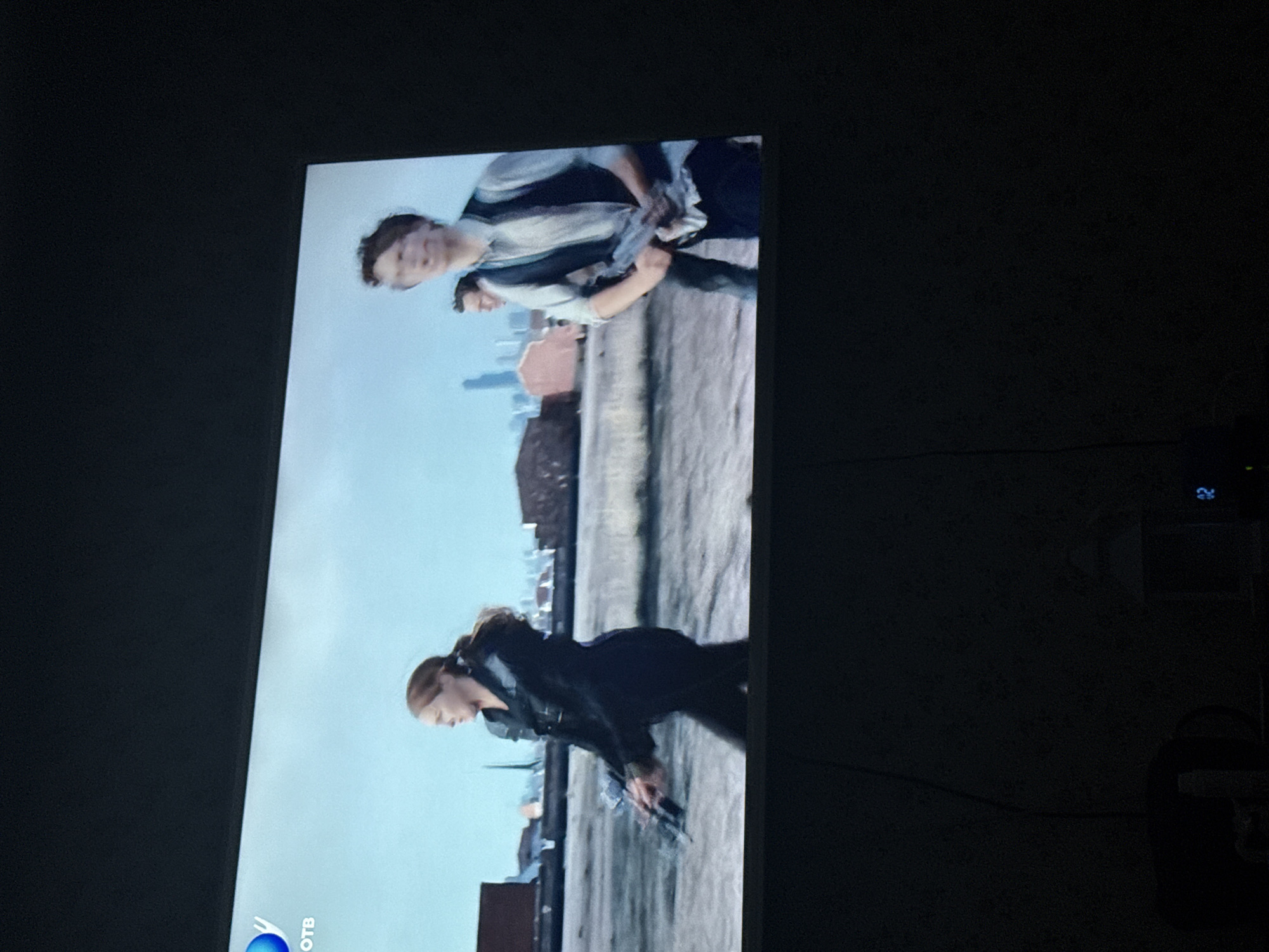 The image shows a scene displayed on a television screen. Two figures stand near the edge of a rooftop, overlooking a city skyline. One appears to be a woman with a ponytail, dressed in dark clothing and holding a gun. The second figure consists of two people standing together, one holding a gun as well. The background features a bright blue sky and a cityscape. The television screen also displays a logo in the corner. The location of Mytishchi, Russia is not discernible.