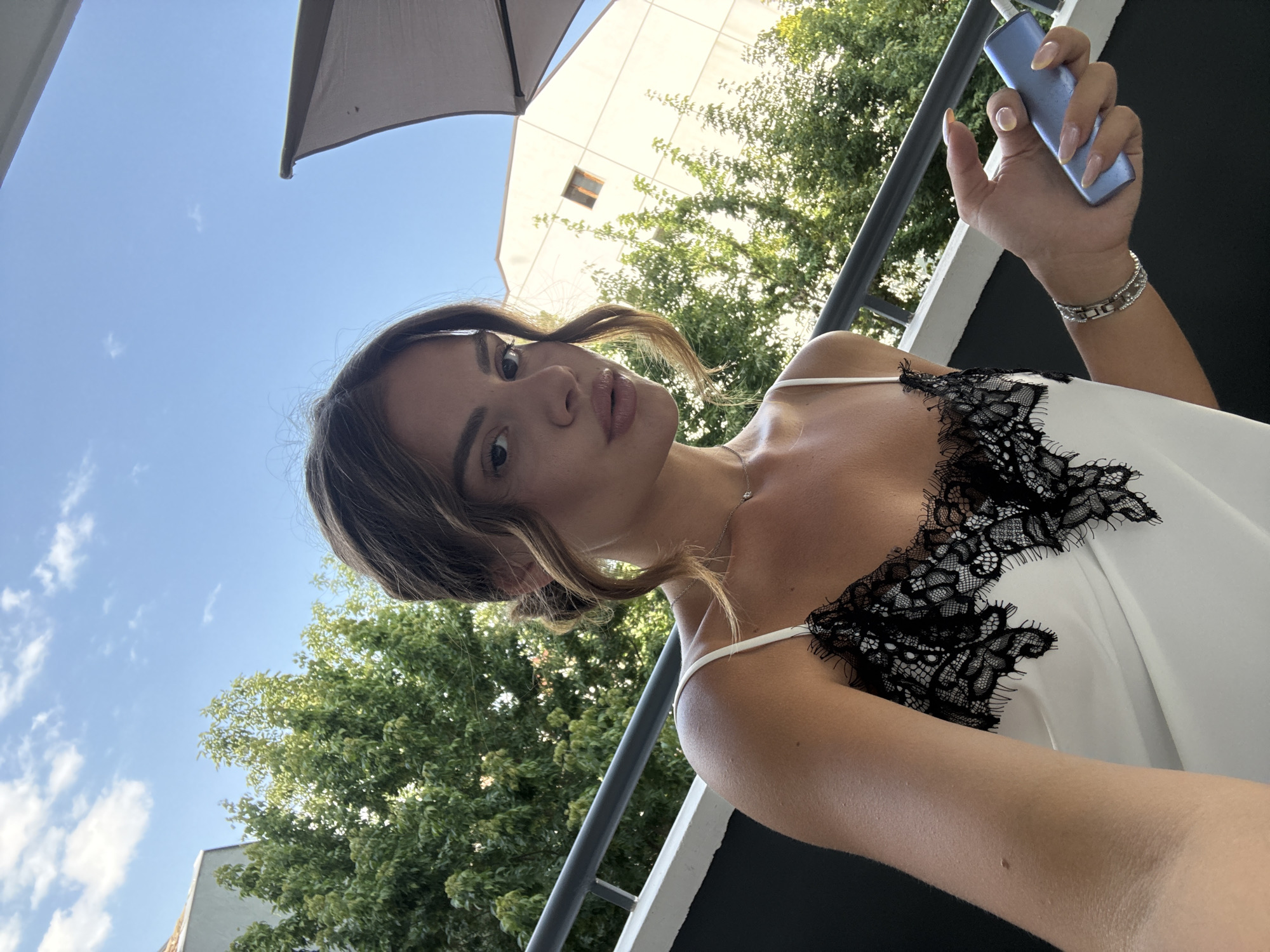A young woman with brown hair styled in an updo is taking a selfie outdoors. She is wearing a white camisole with black lace trim and a delicate necklace. In her right hand, she holds a blue electronic device. She is standing on a balcony with a railing, and behind her are trees, buildings, and a blue sky with clouds. The scene is set during the daytime. The architectural style of the building in the background, with its light-colored facade and visible windows, is consistent with buildings found in cities like Plovdiv, Bulgaria, particularly in residential or commercial areas.
