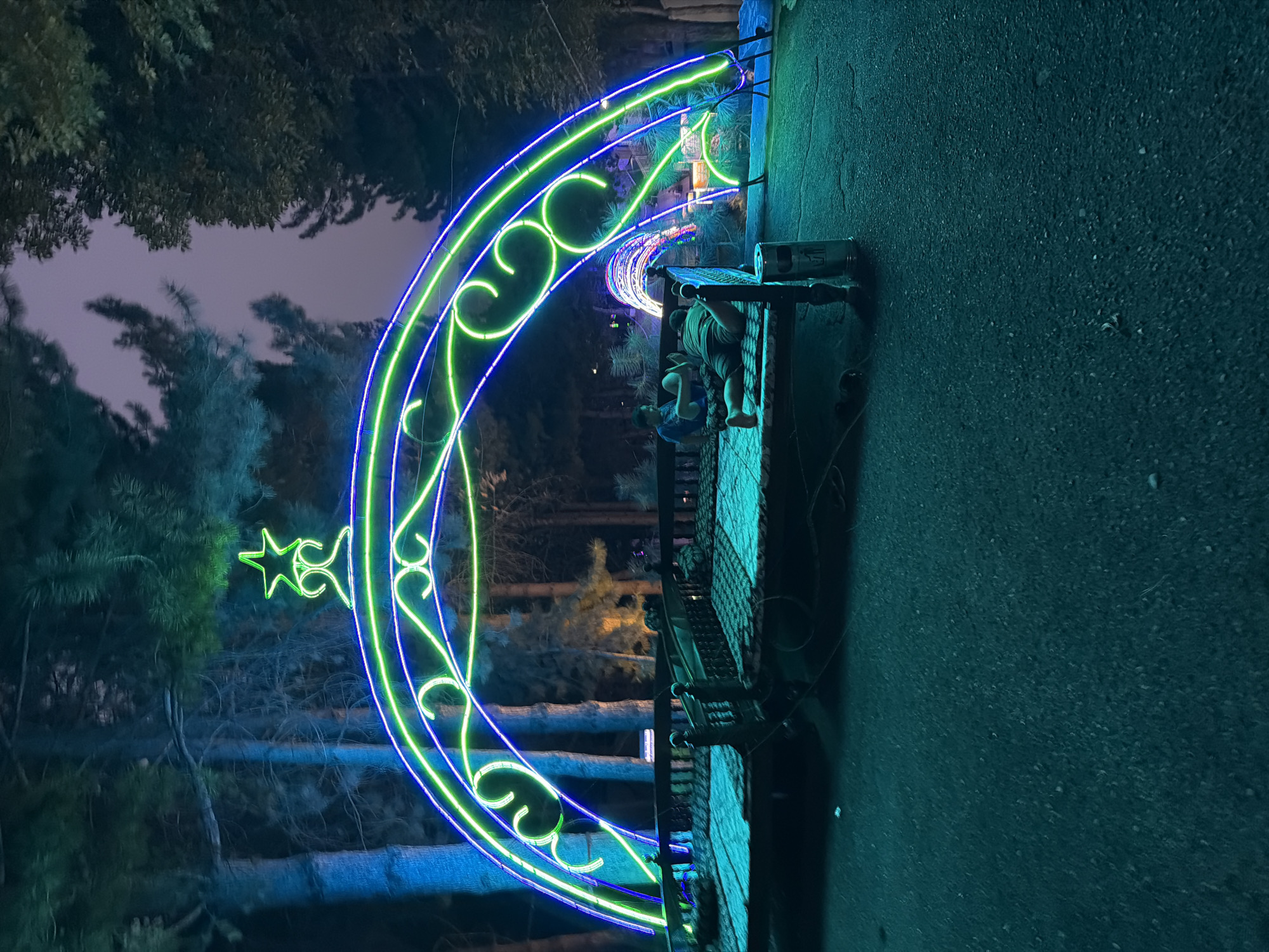 The image depicts an outdoor scene at night, likely a park or public area, illuminated by vibrant neon lights. A large, arched structure made of blue and green neon lights, topped with a star, dominates the foreground. Beneath this arch is a raised wooden platform with railings, resembling a bridge or walkway. On this platform, two individuals are visible. One person is lying down on their stomach, facing away from the camera, appearing relaxed. Another person is seated, facing towards the right side of the frame.
In the background, more neon lights form a tunnel-like structure, extending into the darkness. The environment is surrounded by trees, with some illuminated by the blue neon lights, giving them an ethereal glow. The ground in the foreground is asphalt, with a teal-green hue reflecting the surrounding lights. The overall atmosphere suggests a festive or decorative lighting installation, possibly for a holiday or special event. The location is identified as the city of Khumsan, Uzbekistan.