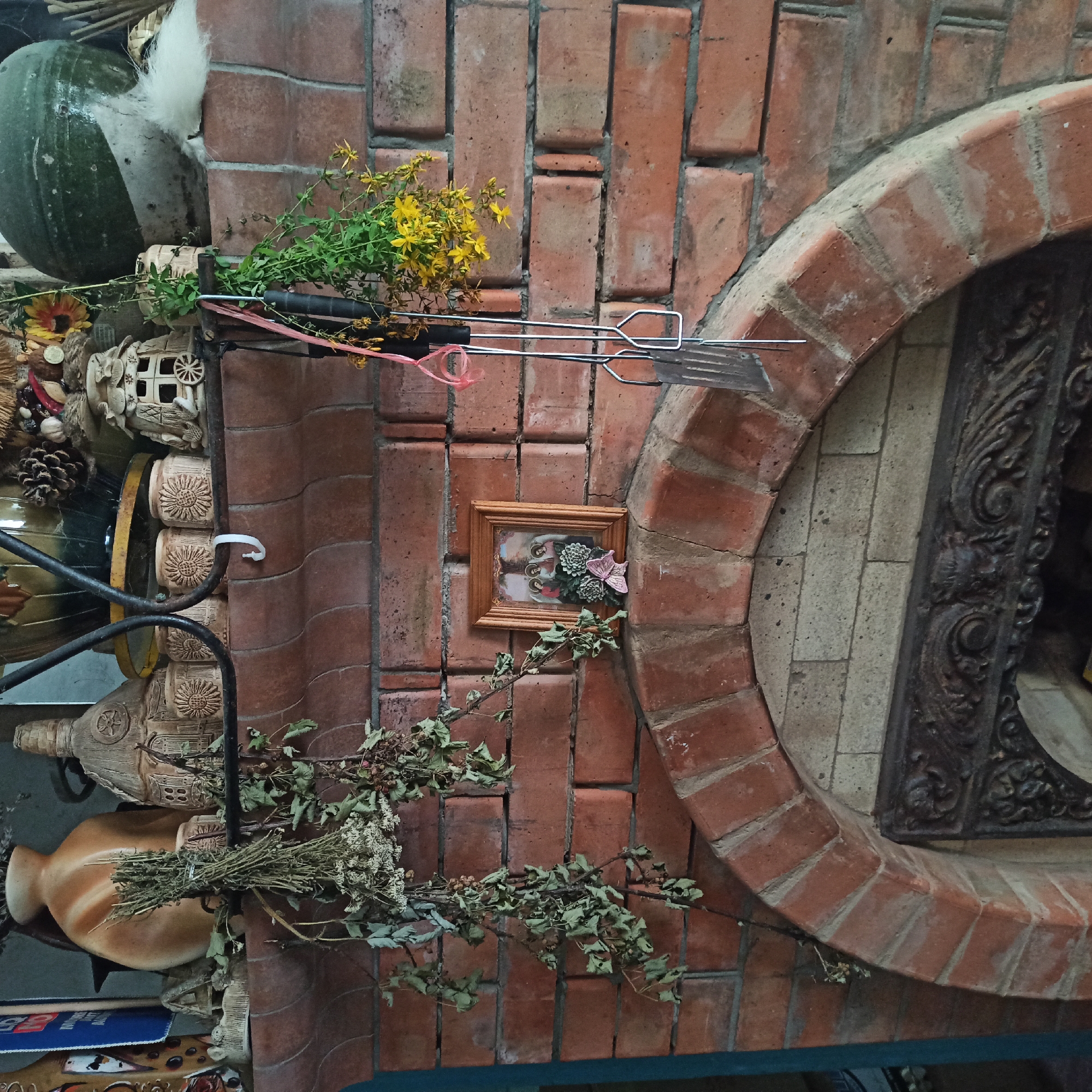 An outdoor brick fireplace, in an unknown location, is decorated with various items. A framed picture of three figures with halos is placed on a shelf above the fireplace opening. To the left of the picture, a bundle of dried herbs hangs down. To the right, a bouquet of yellow flowers and green leaves is attached to a set of grilling utensils. The fireplace has an arched opening with decorative metalwork. The surrounding brickwork is rough and rustic, suggesting an aged or handmade construction.