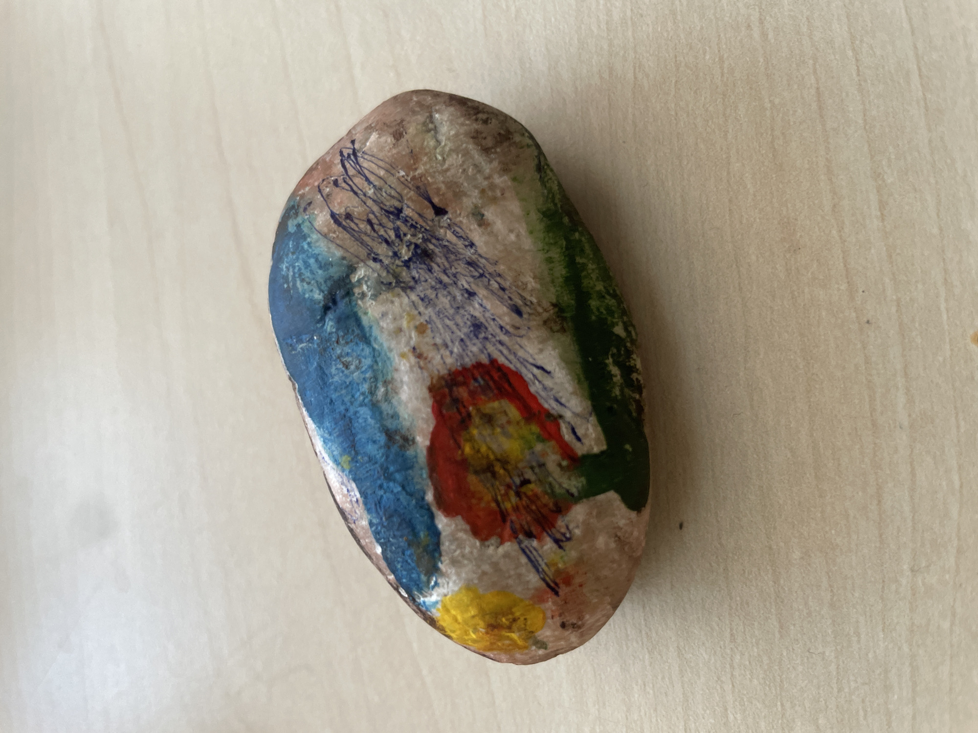 A painted rock rests on a wooden surface. The rock is oval-shaped and features abstract artwork in blue, yellow, red, green, and black. The blue paint appears on the upper left, with yellow and red splotches below it. Streaks of blue marker or ink are visible on the right side of the rock, with green paint along the bottom edge. The wooden surface is light brown with a visible wood grain pattern. The image appears to be taken from a high-angle shot, with the rock positioned slightly off-center. No people or specific city landmarks are visible.