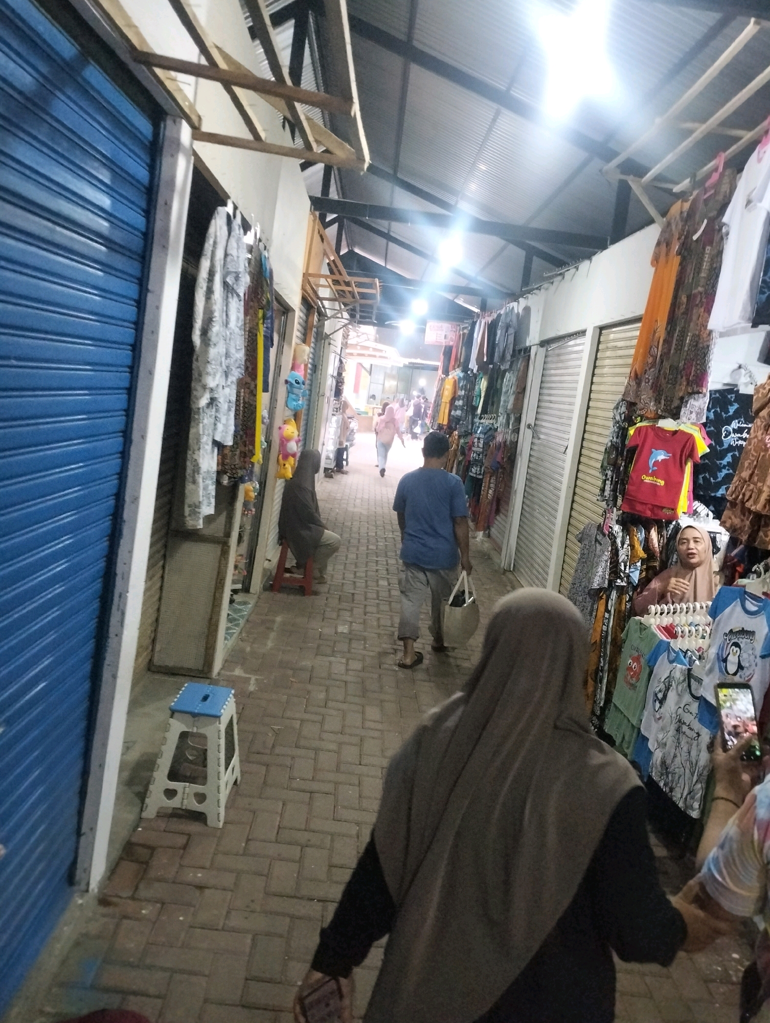 The image depicts a bustling marketplace in Karangbanjar, Indonesia, with stalls lining a narrow walkway. On the left, a blue roller shutter door suggests a closed shop, while adjacent stalls display a variety of clothing items hanging from racks. The walkway is paved with brick-like tiles.
In the foreground, a person wearing a grey hijab and dark clothing is seen from behind, holding a phone. To their right, a woman in a hijab is attending to a stall displaying colorful t-shirts. Further down the walkway, a man in a blue t-shirt and grey pants walks away from the camera, carrying a white bag. Another person in a dark hooded garment sits on a red stool on the left side of the walkway.
The marketplace appears to be covered by a corrugated metal roof supported by wooden beams. Bright lights illuminate the scene, indicating it might be evening or the market is indoors. The stalls are filled with merchandise, primarily clothing, including t-shirts with various graphics, and what appear to be dresses or skirts. Small stuffed toys are also visible in one of the stalls. The overall atmosphere suggests a lively commercial environment.