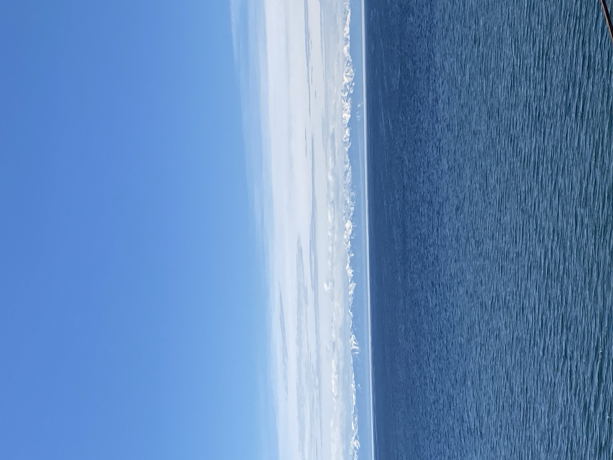 The image shows a view of Lake Baikal from Listvyanka, Russia. The right side of the image is dominated by the dark blue water of the lake, with visible ripples and texture on the surface. To the left of the lake, a thin strip of land is visible, with snow-capped mountains in the distance. The left side of the image is occupied by a clear blue sky with wispy clouds stretching towards the mountains.