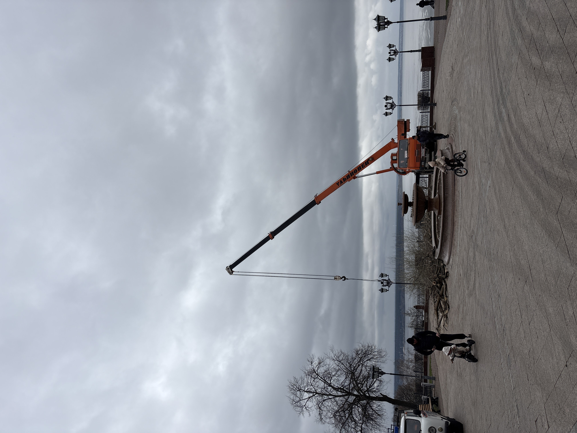 The image shows a cloudy day on a paved area near the Volga River in Ulyanovsk, Russia. An orange "УЛЬЯНОВЕЦ" crane is positioned next to a fountain. Two adults are present; one is helping a child ride a small bicycle. Nearby, there are cut branches on the ground. A vehicle is parked near a bare tree, with streetlights lining the embankment.