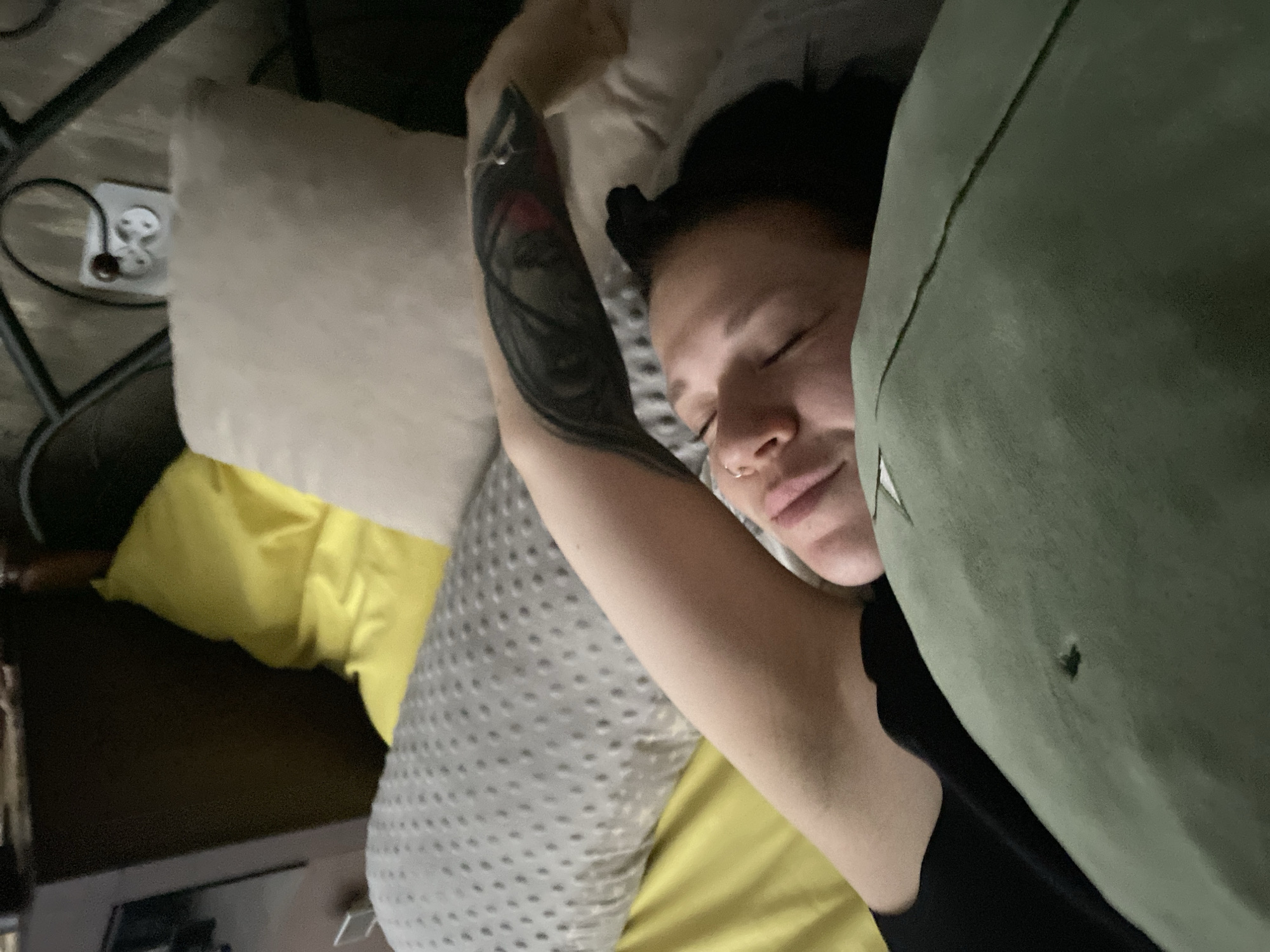 A woman with black hair pulled back in two buns sleeps in a bed with an olive-green duvet pulled up to her face. Her left arm is raised, revealing a tattoo. Pillows of varying colors (white, grey with polka dots, yellow) surround her. A black metal bed frame and electrical outlet are visible in the background. The location is inferred to be Chaikovsky, Russia, based on prior knowledge, though there is no explicit confirmation in the image itself.