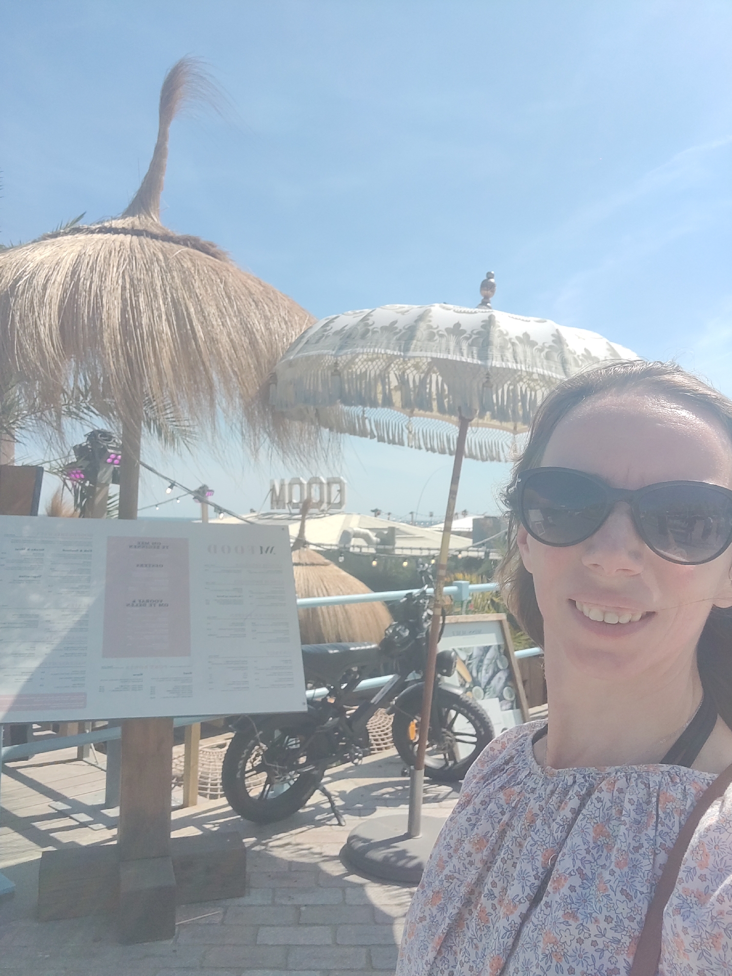 A woman with brown hair and sunglasses smiles at the camera in a selfie. She is wearing a floral print blouse. Behind her, a thatched umbrella and a lighter-colored umbrella are visible. A sign that reads "MOOD" is in the background, along with a motorcycle and a menu. The scene appears to be an outdoor cafe or bar, possibly in The Hague, Netherlands, given the architecture and ambiance. The lighting suggests it is daytime.