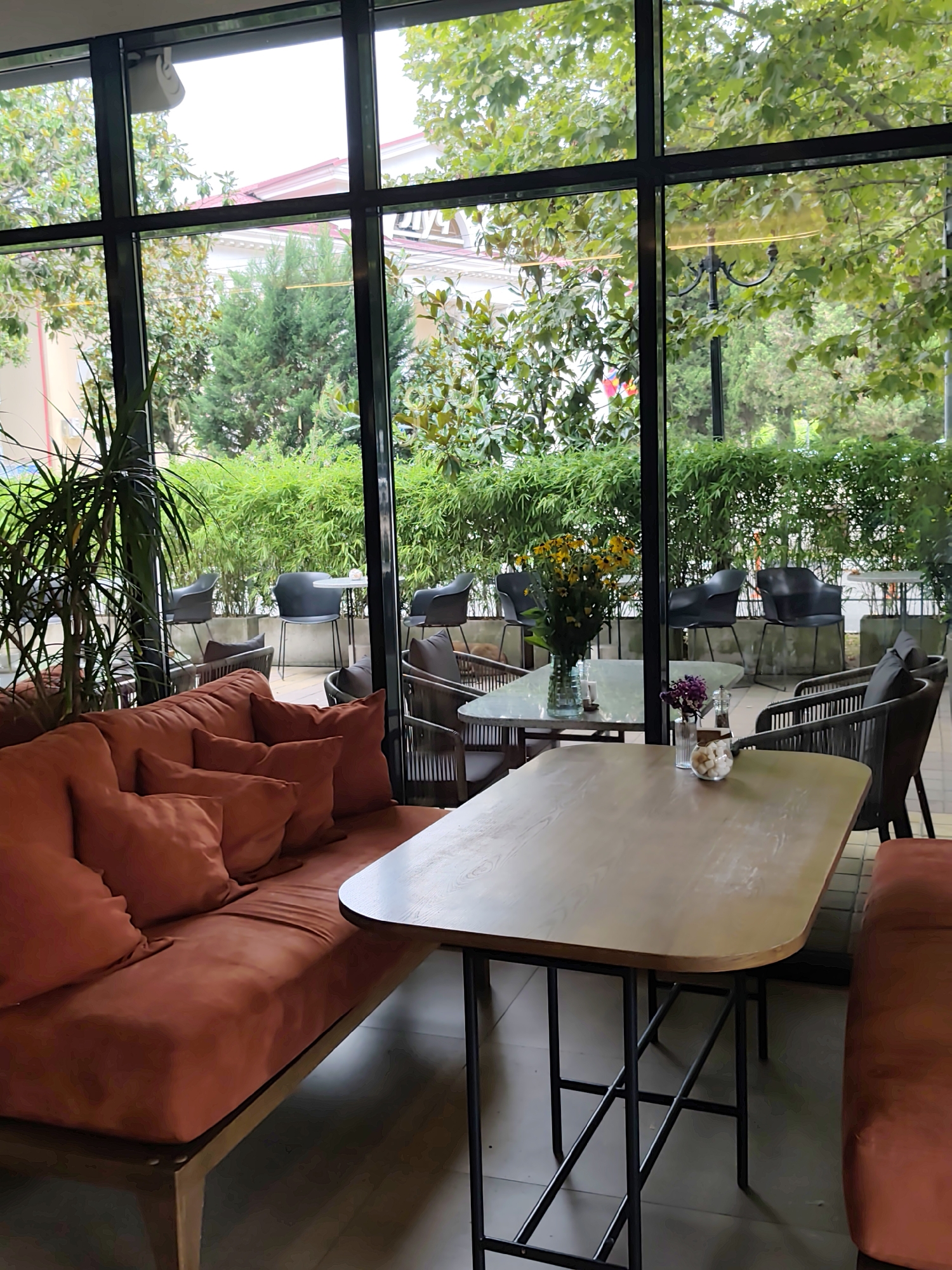 The image depicts the interior of a cafe or restaurant with large windows overlooking a lush green outdoor area. The cafe features modern, comfortable seating, including an orange sectional sofa with plush cushions and several dark-colored chairs around tables. One table in the foreground is a light-colored wooden rectangle with a black metal frame. A glass vase filled with yellow flowers sits on a marble-topped table in the background, along with another smaller vase containing purple flowers and some decorative white pebbles. Outside, a dense hedge or bamboo-like plants form a backdrop, with trees and buildings visible beyond. The overall ambiance suggests a tranquil setting. The presence of this cafe with a view of greenery aligns with the characteristic offerings found in resort towns like Sochi, Russia, known for their natural beauty and recreational facilities, often incorporating outdoor seating or large windows to maximize views.