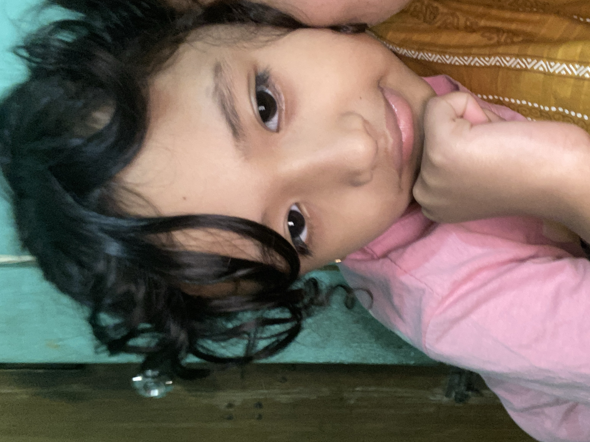 A young girl with dark, curly hair is posing for a close-up photograph. She is wearing a pink t-shirt and has her right hand resting on her chin. Her left hand is also visible and appears to be resting on her chest. She is looking directly at the camera with a neutral expression. The background is a teal-colored wall and a wooden doorframe. The image appears to have been taken indoors in a residential setting in Dhaka, Bangladesh.