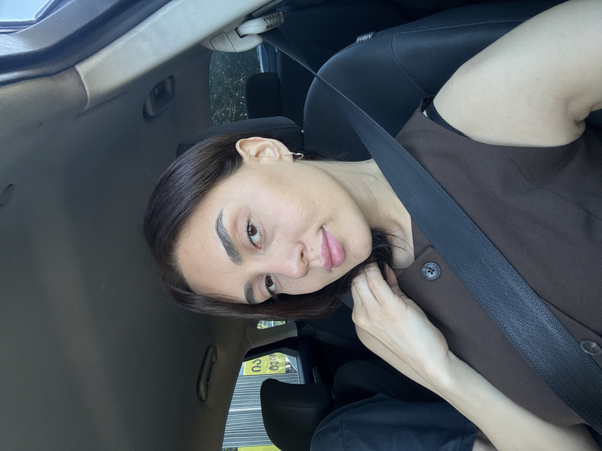 A young woman with dark hair and light skin is seen from the chest up, sitting in the driver's seat of a car. She is wearing a brown sleeveless top with two dark buttons on the front. A black seatbelt is diagonally across her chest. Her right hand is touching her neck. She has a small silver heart-shaped earring in her left ear. In the background, through the car window, a yellow sign with Georgian writing is visible, suggesting the location is Tbilisi, Georgia. The car interior is visible, including the car's headrest and a portion of the passenger seat.