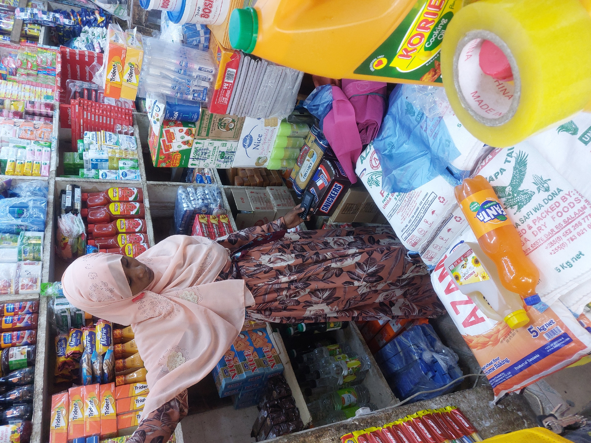 The image shows a person wearing a peach-colored hijab and floral-patterned dress, likely shopping in a small convenience store in Dar es Salaam, Tanzania. The store is densely packed with various consumer goods including food items, toiletries, and household supplies.
Specific items visible include packets of "Trident" gum, bottles of "Korie" cooking oil, boxes of "Nice" biscuits, "Fanta" soda, bags of "Azam" flour, "Snickers" bars, and other packaged snacks and groceries. Brands like "Whitedent", "Protex", and "Marie" biscuits are also visible.
The store appears to be a traditional, possibly family-run, retail operation, typical in many parts of Dar es Salaam. The goods are stacked high on shelves and in bins, maximizing the use of space. A roll of yellow tape sits atop the merchandise, suggesting a point-of-sale activity. The bag of "Apa Taifa" flour has text indicating it's packed by S.A. DRY FOODS in Dar es Salaam, further pinpointing the location.