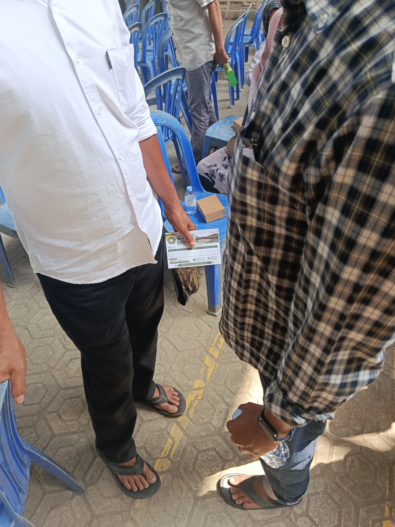 In Kawunganten Lor, Indonesia, two individuals stand in what appears to be an outdoor public gathering. The person on the left wears a white long-sleeved shirt and black pants, holding a small card or pamphlet. The person on the right is dressed in a plaid long-sleeved shirt and blue jeans. They are holding a bottle of water. The background shows rows of blue plastic chairs and other attendees in the distance. The ground is paved with hexagonal tiles and marked with yellow lines. The lighting suggests it is daytime with bright sunlight.