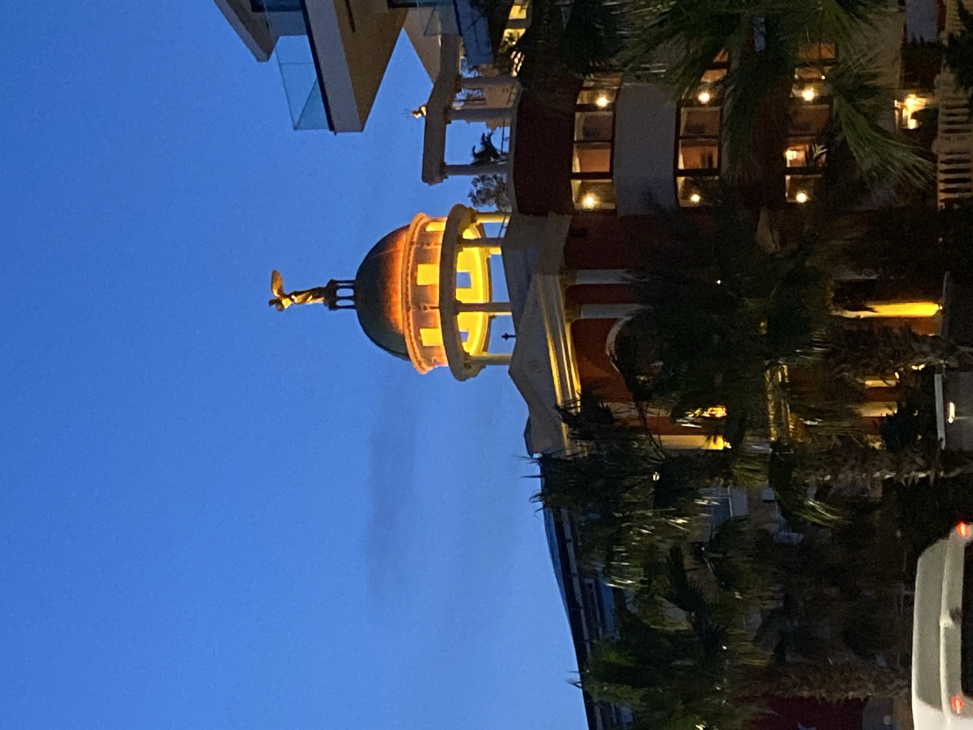 Here is a detailed description of the image:
The image shows a building at night in Alanya, Turkey. The building features a dome with an angel statue on top. The dome is illuminated from within. The building also has windows with lights visible inside. Palm trees surround the building. The sky is dark blue.