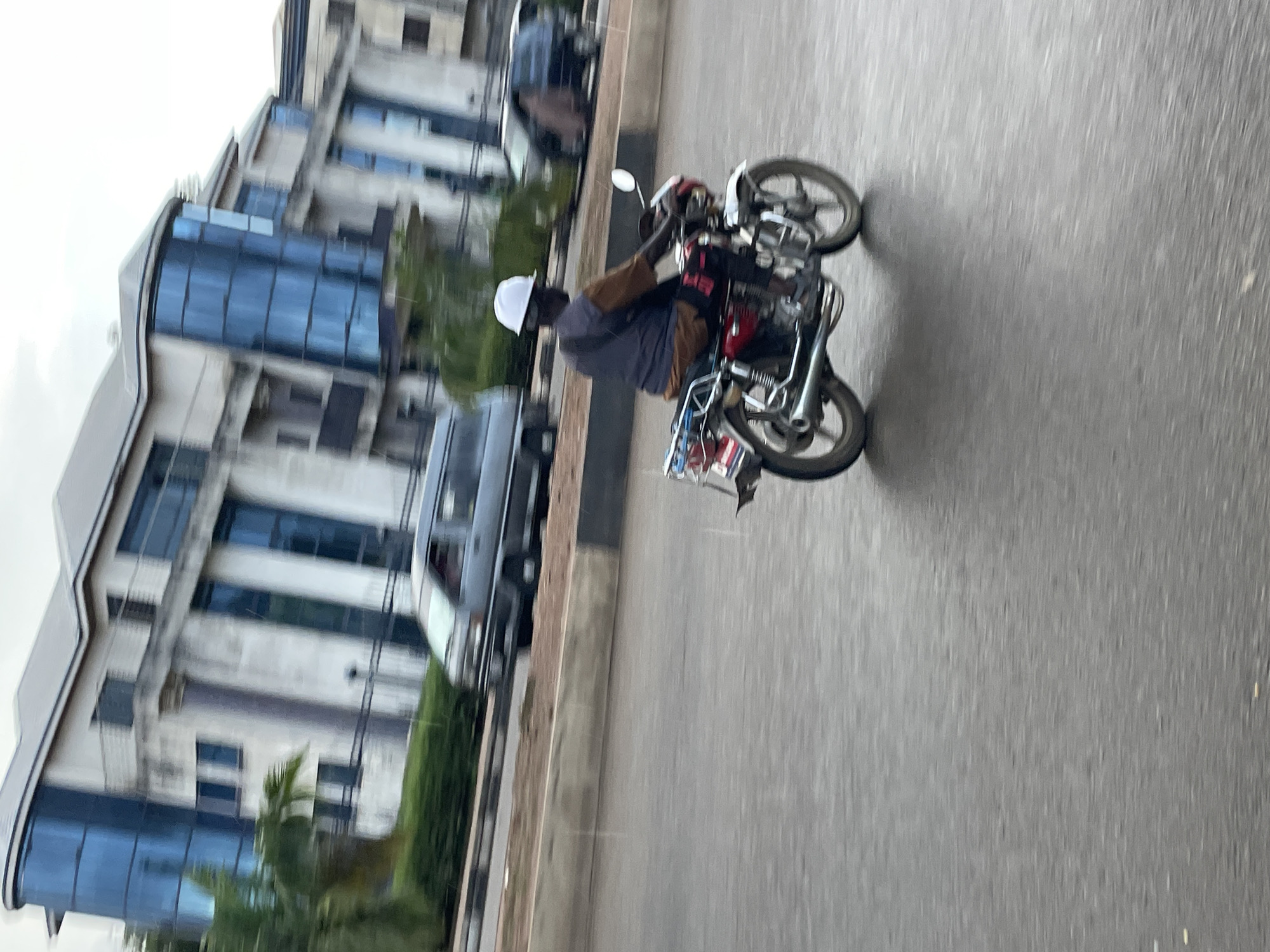 The image depicts a street scene in Jos, Nigeria, during what appears to be overcast or rainy weather, suggested by the wet asphalt and visible raindrops. In the foreground, a person is riding a red motorcycle. The rider is wearing a white helmet and a blue vest over a brown shirt. The motorcycle has a chrome exhaust pipe and a luggage rack on the back. In the middle ground, a silver van is driving on the road. Behind the van, a large, modern building with blue-tinted windows and white siding is visible. The building has multiple floors and balconies. The overall impression is one of urban activity in a tropical environment.