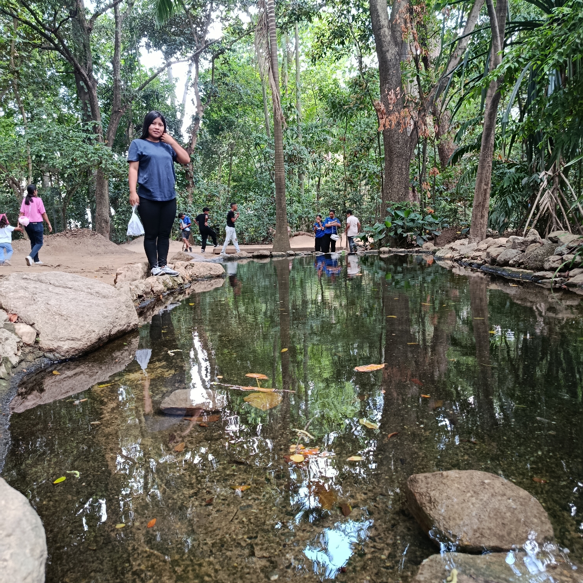 The image shows a natural setting, likely a park or a botanical garden. A pond occupies the center of the frame, reflecting the surrounding trees. A young woman stands on the edge of the pond, wearing a blue t-shirt and dark pants, holding a white plastic bag. Other people can be seen in the background, some standing by the pond's edge and others walking along a path. The environment is lush with trees and vegetation, creating a serene atmosphere. The location appears to be in an Unknown city.