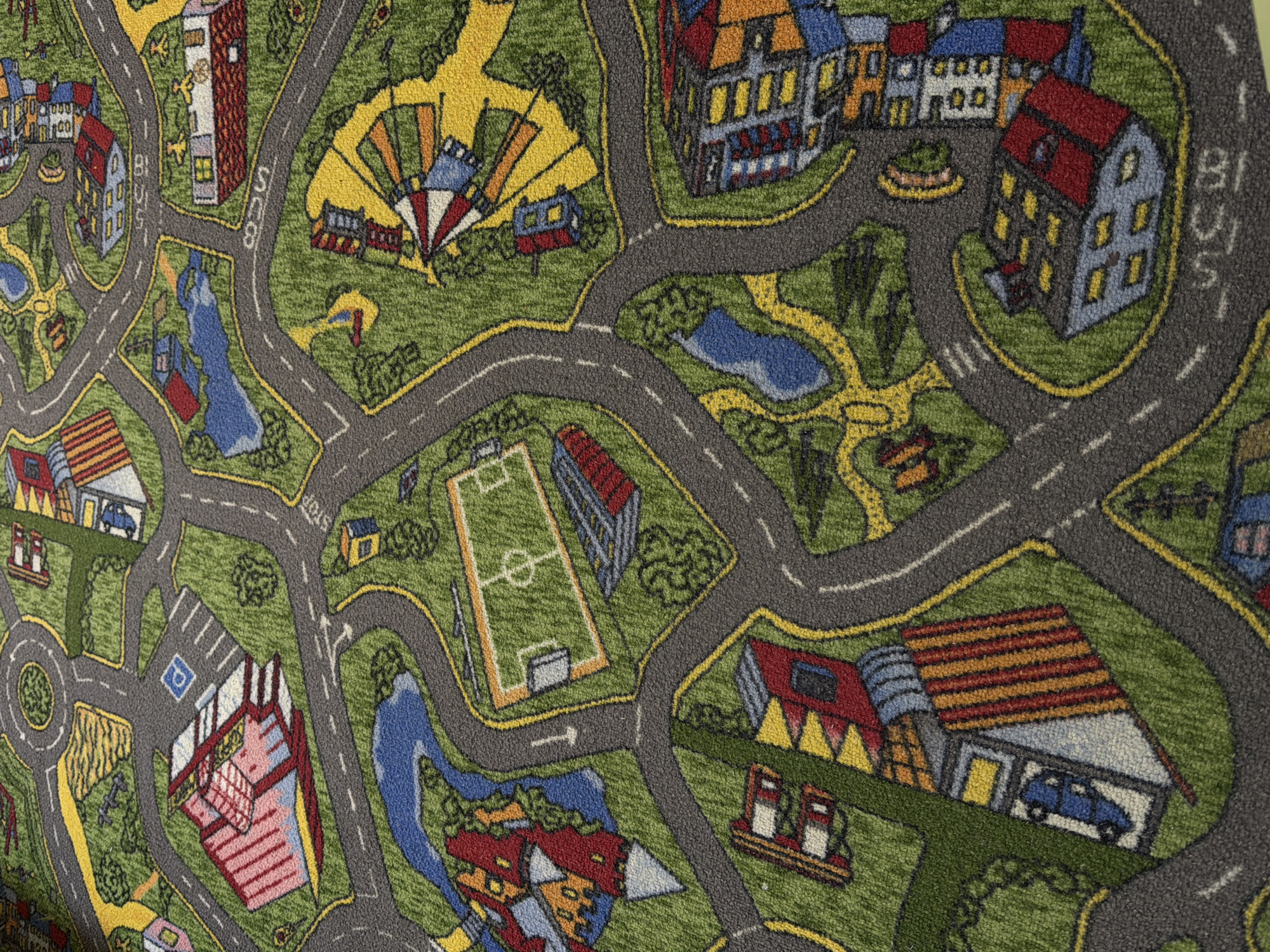 The image is of a children's play rug designed to resemble a miniature city. It includes streets, buildings, a roundabout, a parking lot with a parking symbol, a soccer field, a river, trees, and various structures that mimic houses, schools, and other city facilities. There are representations of cars on the rug. The style is cartoonish and simplistic. The rug's location is not depicted, and there is no information about people or actions. The city of Chaikovsky, Russia, is not depicted in any discernable manner.