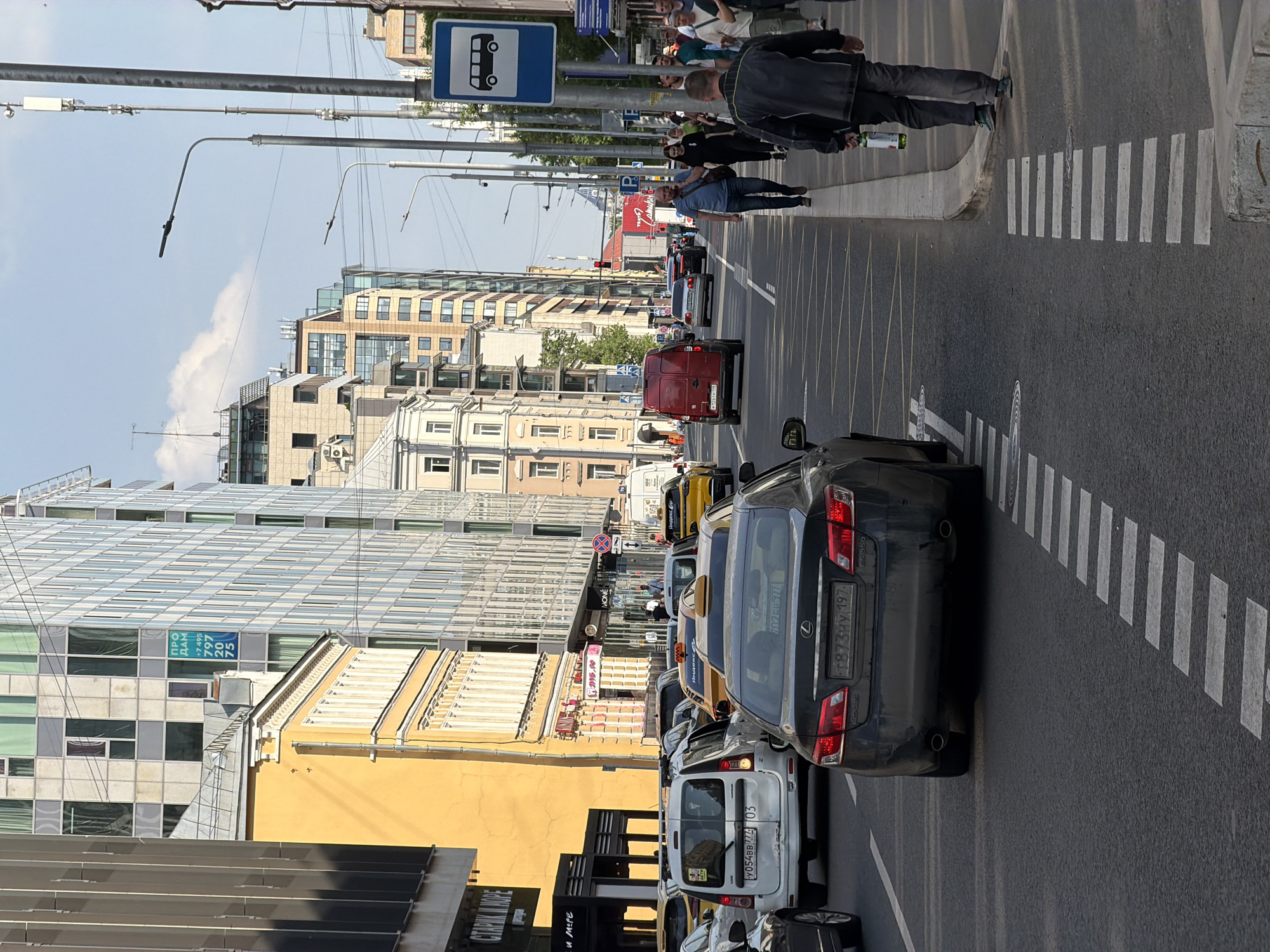 The image depicts a street scene in Moscow, Russia. The street is filled with cars, primarily taxis and vans, indicating traffic congestion. Pedestrians are visible on the sidewalk to the right, with one man carrying a bottle. A bus stop sign is also visible on the right side of the road. Buildings line the street, including modern glass-fronted structures and older, yellow-painted buildings. The license plate numbers and signs are in Russian, further confirming the location.