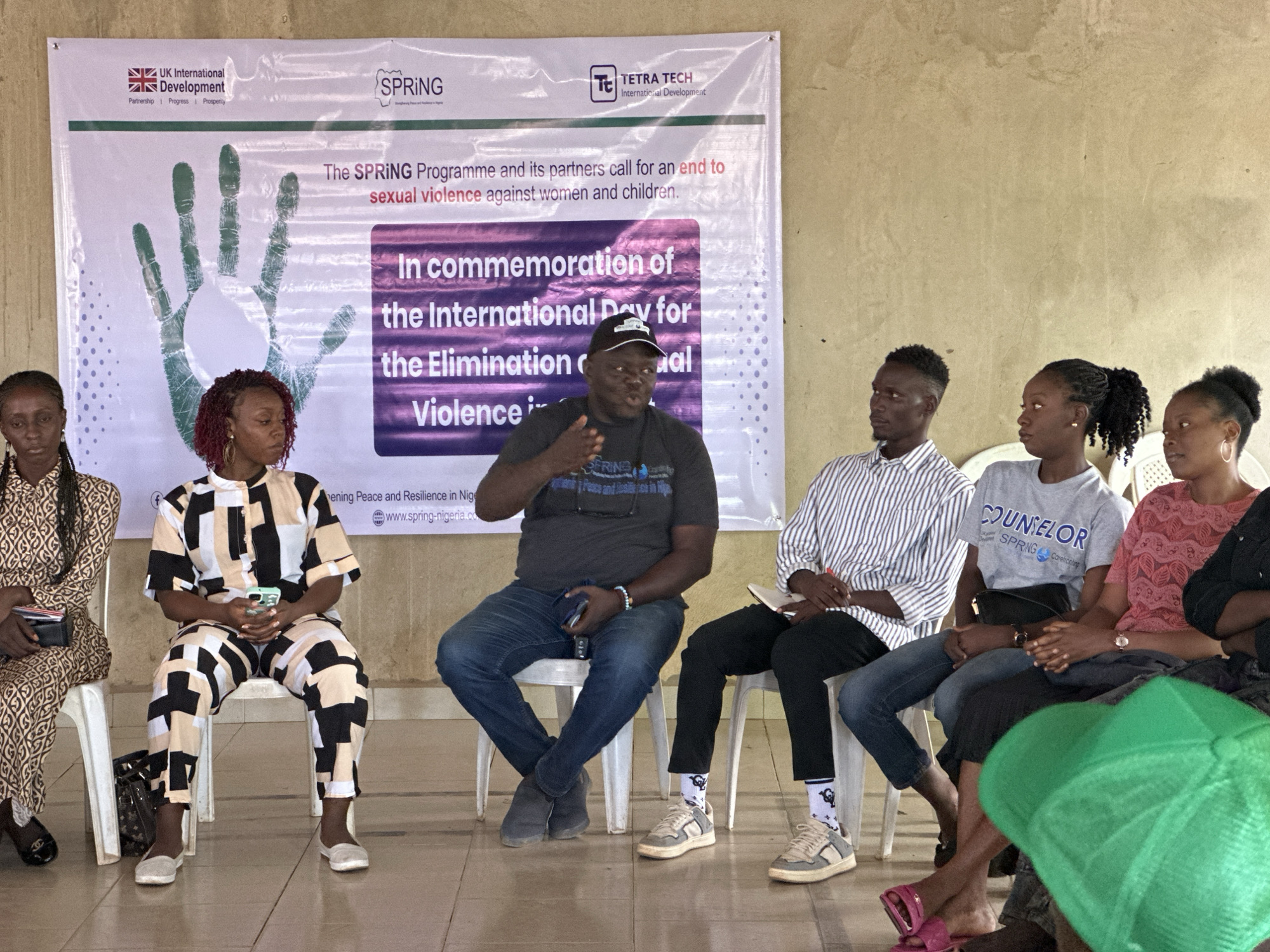 The image depicts a panel discussion or presentation taking place indoors, with a banner in the background that reads "The SPRING Programme and its partners call for an end to sexual violence against women and children. In commemoration of the International Day for the Elimination of Violence". The banner also features logos for "UK International Development", "SPRING", and "TETRA TECH International Development". The setting appears to be in Sabon Tasha, Nigeria, as indicated by the presence of the banner and the context of SPRING's work in the region.
There are six individuals seated on white plastic chairs in front of the banner. From left to right, the first person is a woman wearing a patterned dress and holding a black wallet. The second is a woman in a black, white, and beige patterned outfit, holding a mobile phone. The third is a man wearing a dark grey t-shirt and jeans, speaking and gesturing with his right hand. He is wearing a dark baseball cap. The fourth is a man in a striped shirt and black pants, holding a notebook and pen. The fifth is a woman in a grey t-shirt with "COUNSELOR" and "SPRING" printed on it. The sixth person is a woman in a pink top. The legs of a seventh person wearing a bright green head covering are visible on the far right. The individuals appear to be engaged in the event, with some looking towards the speaker and others at their notes or phones. The floor is tiled, and the wall behind the banner is a light, textured color. The lighting suggests it is daytime, possibly with artificial lighting contributing to the scene.