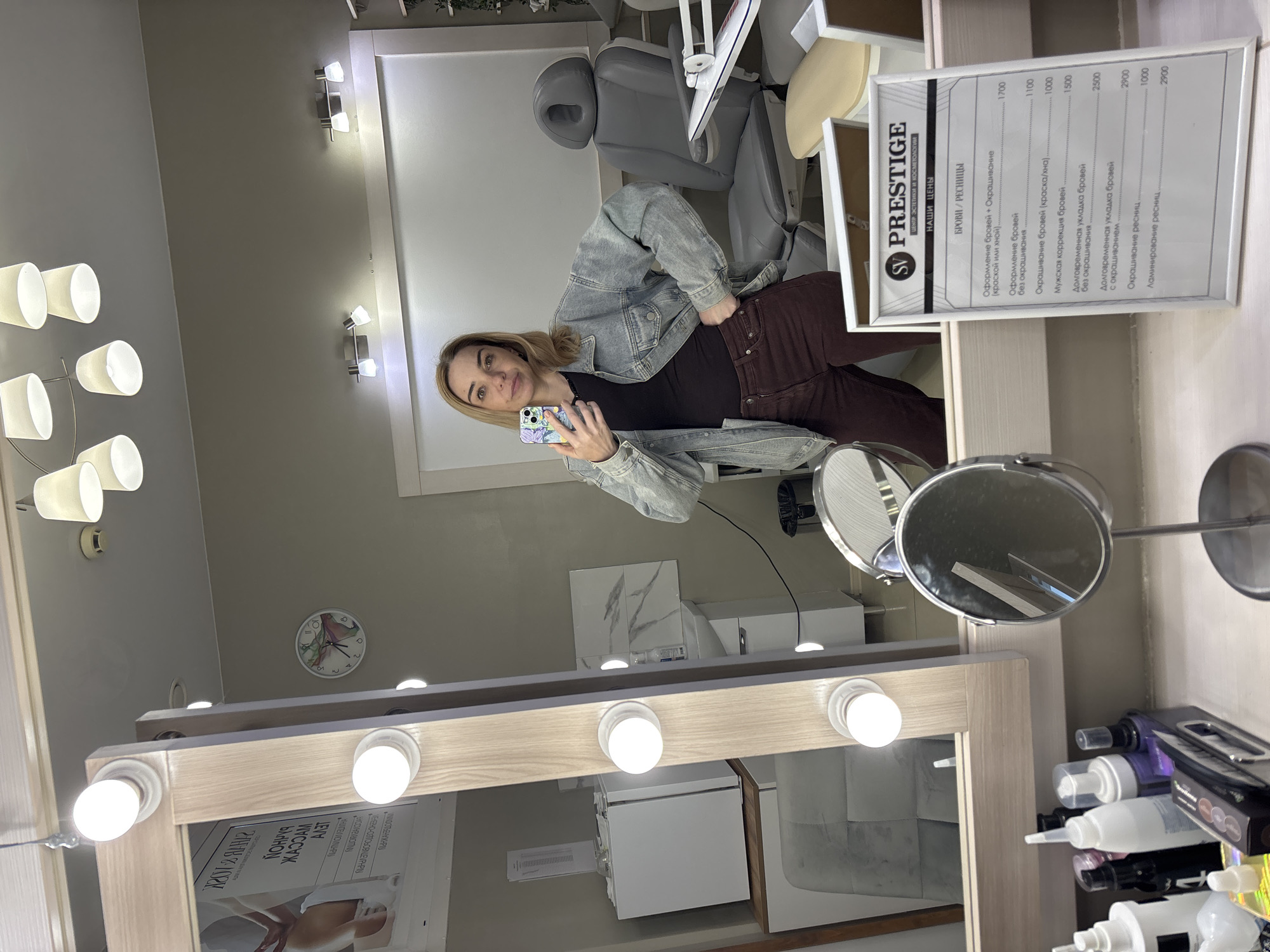 A woman is taking a mirror selfie in a brightly lit room that appears to be a beauty salon. She is wearing a denim jacket over a dark top and maroon pants. She is holding a smartphone in her right hand. To her left is a large vanity mirror with light bulbs around its frame. On the vanity, there are various beauty products and a small, stand-up mirror. Behind her, a grey salon chair is visible, along with a sink and other salon equipment. A price list for "BROWS / LASHES" services is displayed on a white board in the foreground. The overall environment suggests a professional beauty service setting. The location is Ladozhskoe Ozero, Russia.
