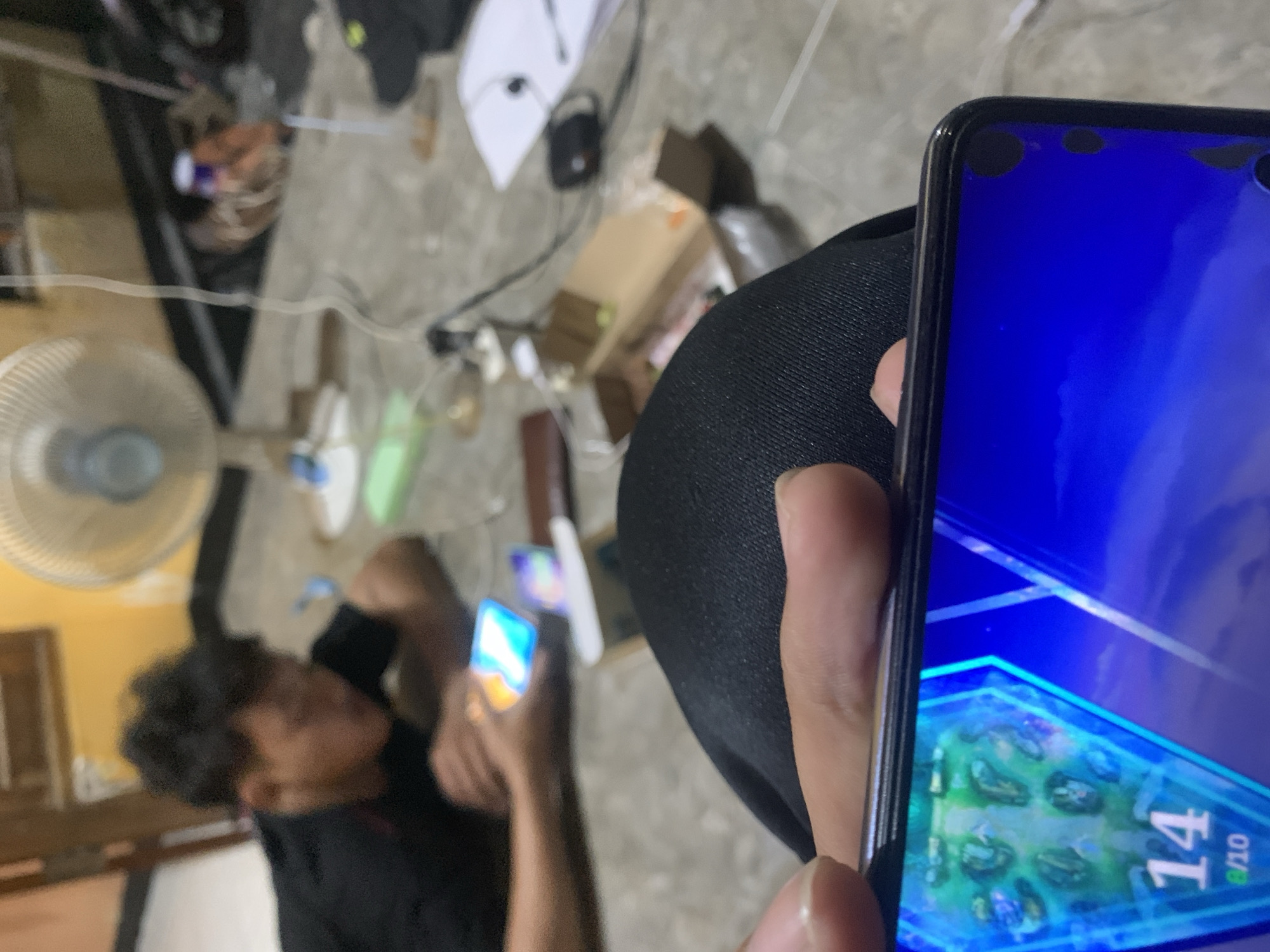The image shows a close-up view of a person holding a smartphone displaying a game with blue and green graphics and the number "14". In the background, a blurred figure wearing a black shirt is visible, also holding a smartphone. The setting appears to be an indoor floor with a fan and various objects scattered around, including a cardboard box and papers. The lighting suggests an indoor environment, possibly a home. The overall scene depicts two individuals engaged with their mobile devices.