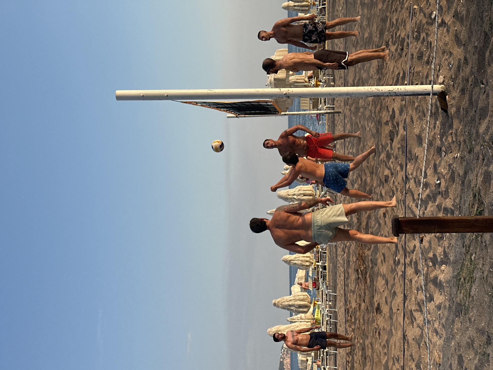 Six men are playing beach volleyball on a sandy beach. The sky is clear and blue. In the background, there are rows of beach chairs and umbrellas. The men are all wearing swim trunks, and they appear to be in good physical condition. One man is in the process of hitting the volleyball, while the others are positioned on the court, ready to receive or play the ball. The game appears to be casual, with the players enjoying themselves. The location is the city of Boreti in Montenegro, a coastal town known for its beaches and clear waters.