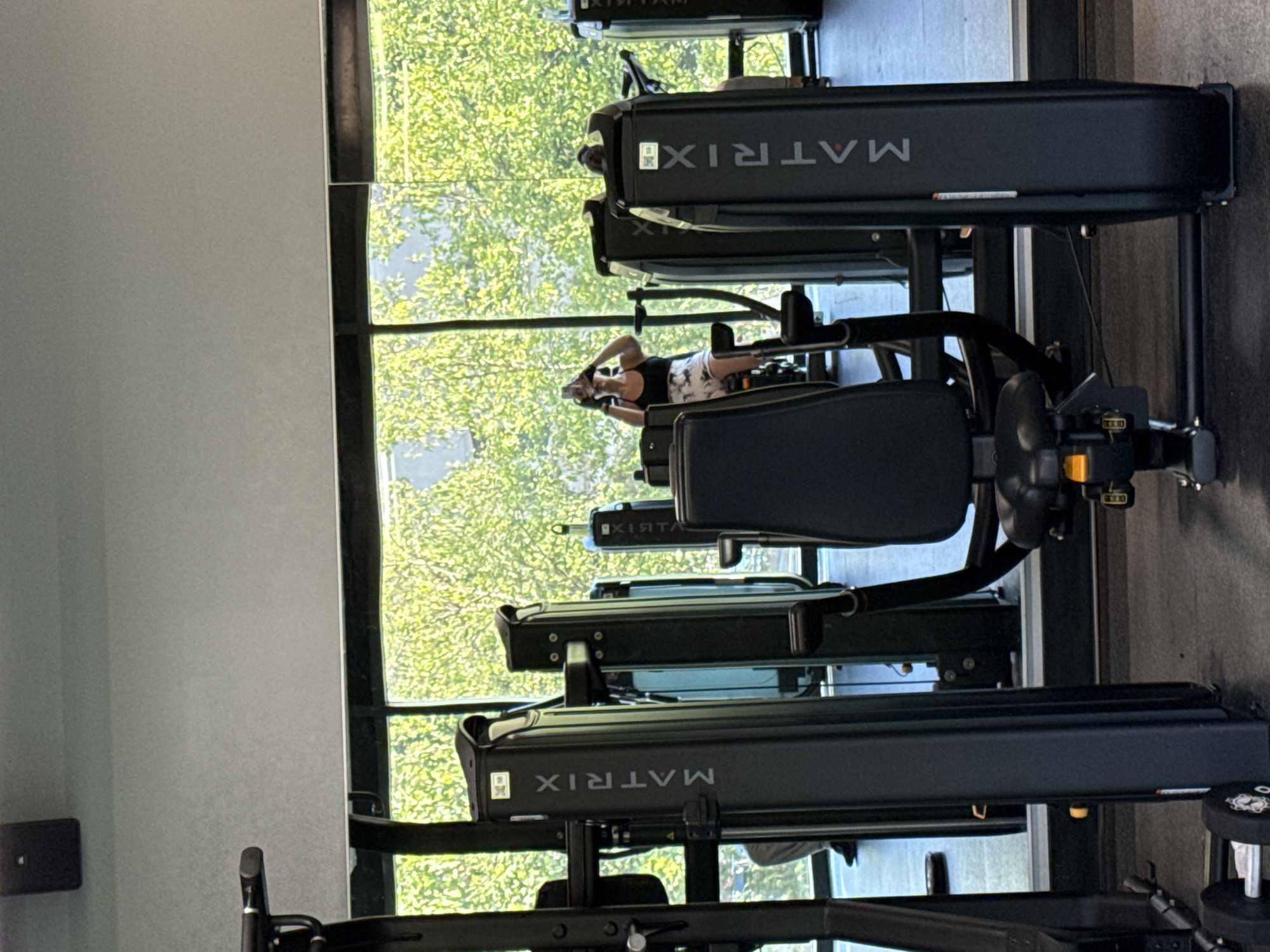 In a gym in the City of Yonkers, United States of America, a person is seen exercising. The person, wearing a black tank top and patterned shorts, is seated on a machine and holding a phone up, likely taking a selfie. Multiple pieces of gym equipment are visible, including a machine with the "MATRIX" logo prominently displayed. The gym features large windows with a view of lush green trees, suggesting an outdoor natural setting beyond the glass. The floor is a dark, gym-style surface, and the overall lighting indicates a daytime scene. The perspective is from within the gym, looking towards the window and the person exercising.