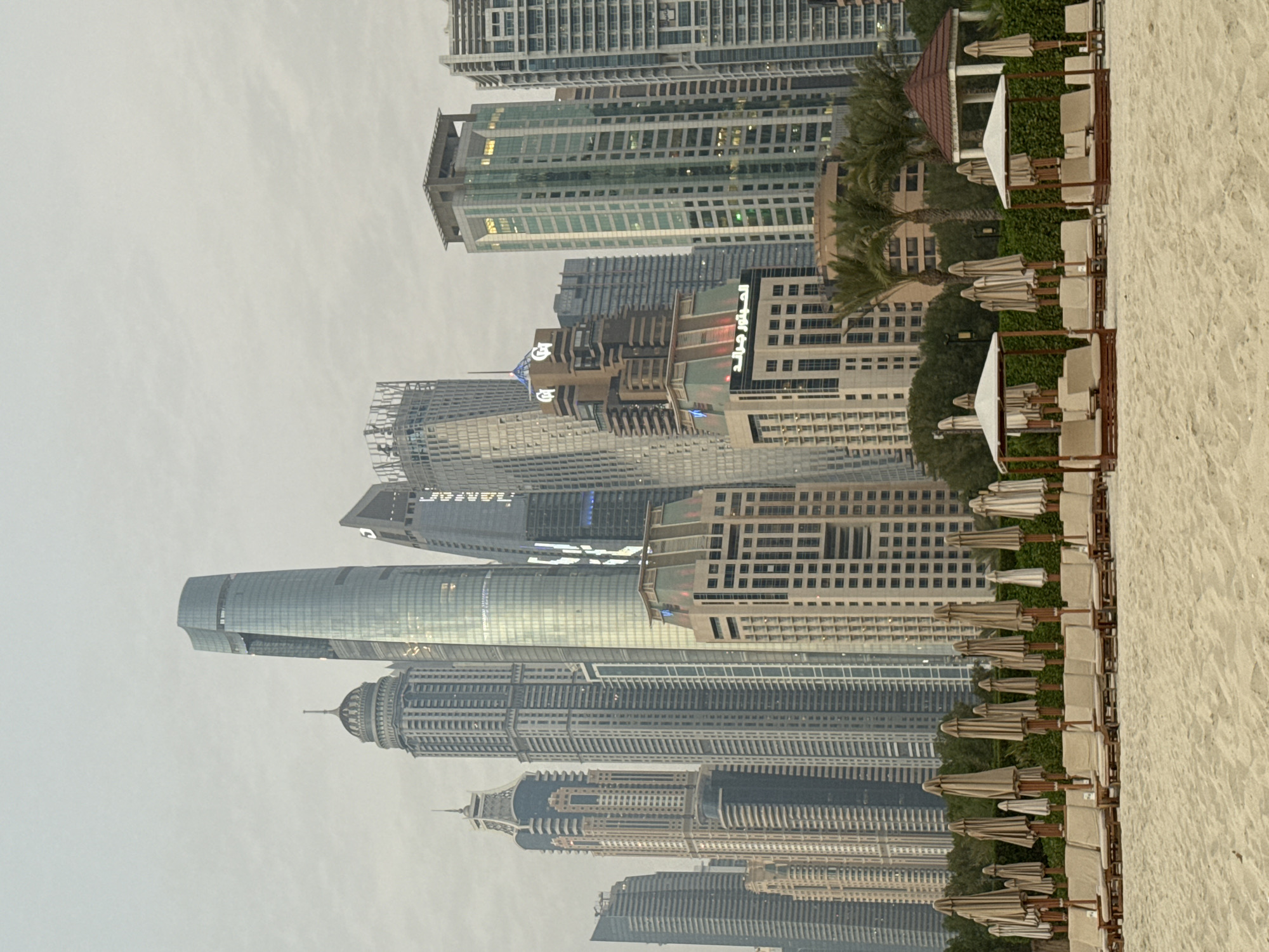 The image depicts a serene beach scene in Dubai, United Arab Emirates, with a backdrop of the city's iconic skyscrapers. The foreground is dominated by a pristine sandy beach, where rows of lounge chairs and umbrellas are neatly arranged, indicating a well-maintained resort or public beach area.
Behind the beach, lush green trees and palm trees create a natural buffer, separating the tranquil beach from the bustling urban landscape. The middle ground features a collection of modern, high-rise buildings that are characteristic of Dubai's renowned skyline. These structures vary in architectural style and height, with some featuring sleek glass facades and intricate designs. One prominent building displays Arabic text, suggesting it is a commercial or residential complex.
The sky above is overcast, lending a soft, diffused light to the scene. The overall atmosphere is one of calm and tranquility, with the juxtaposition of the natural beach environment and the man-made architectural marvels of the city. There are no people visible in the image, allowing the viewer to focus on the interplay between nature and urban development. The image appears to be a photograph taken during daylight hours.