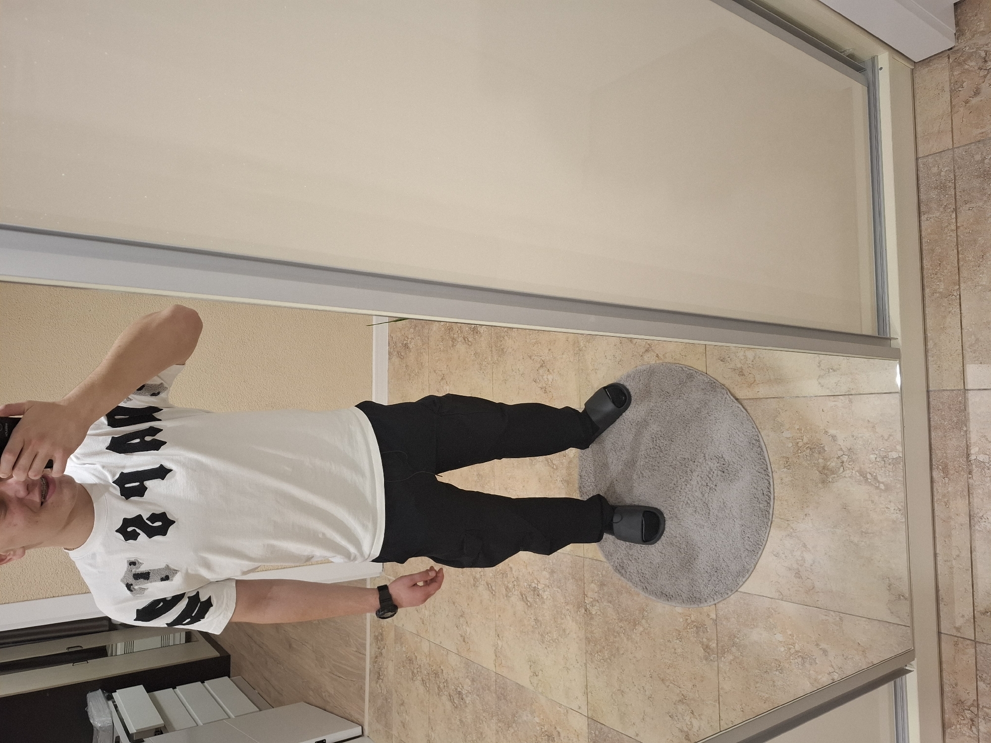 A person is standing on a light-colored, round bath mat in a bathroom. The person is wearing a white t-shirt with large, black gothic-style lettering, black pants, and black slide sandals. They are holding a phone and taking a picture in a mirror. The bathroom has beige tile flooring, and a sliding glass shower door is visible. It is not possible to determine if the location is in Veliki Preslav, Bulgaria.