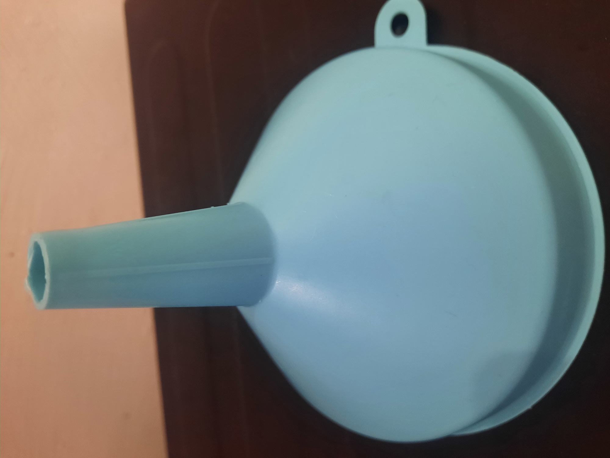 A light blue plastic funnel is positioned on a dark brown surface, which appears to be a table. The funnel is oriented vertically, with its conical section widening towards the bottom and its cylindrical tube extending upwards. The background consists of a peachy-beige wall. The image is a close-up shot, focusing entirely on the funnel and its immediate surroundings. There are no people or animals present, and no actions are being depicted. The environment is static and simple. The location in Lhokseumawe, Indonesia, is not discernible from the visual information provided in the image.